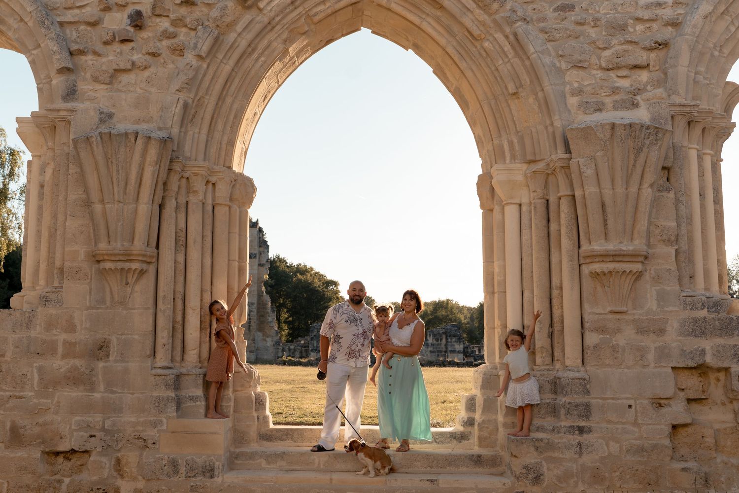 A family poses under a stone arch at historic ruins during golden hour with their dog.