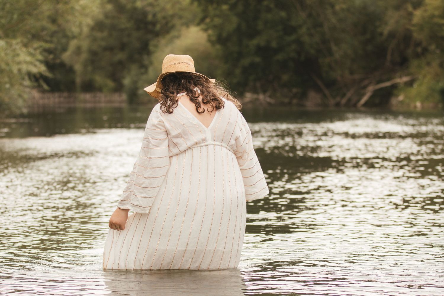 Pregnant woman in white dress and straw hat wading in a river surrounded by lush green trees.
