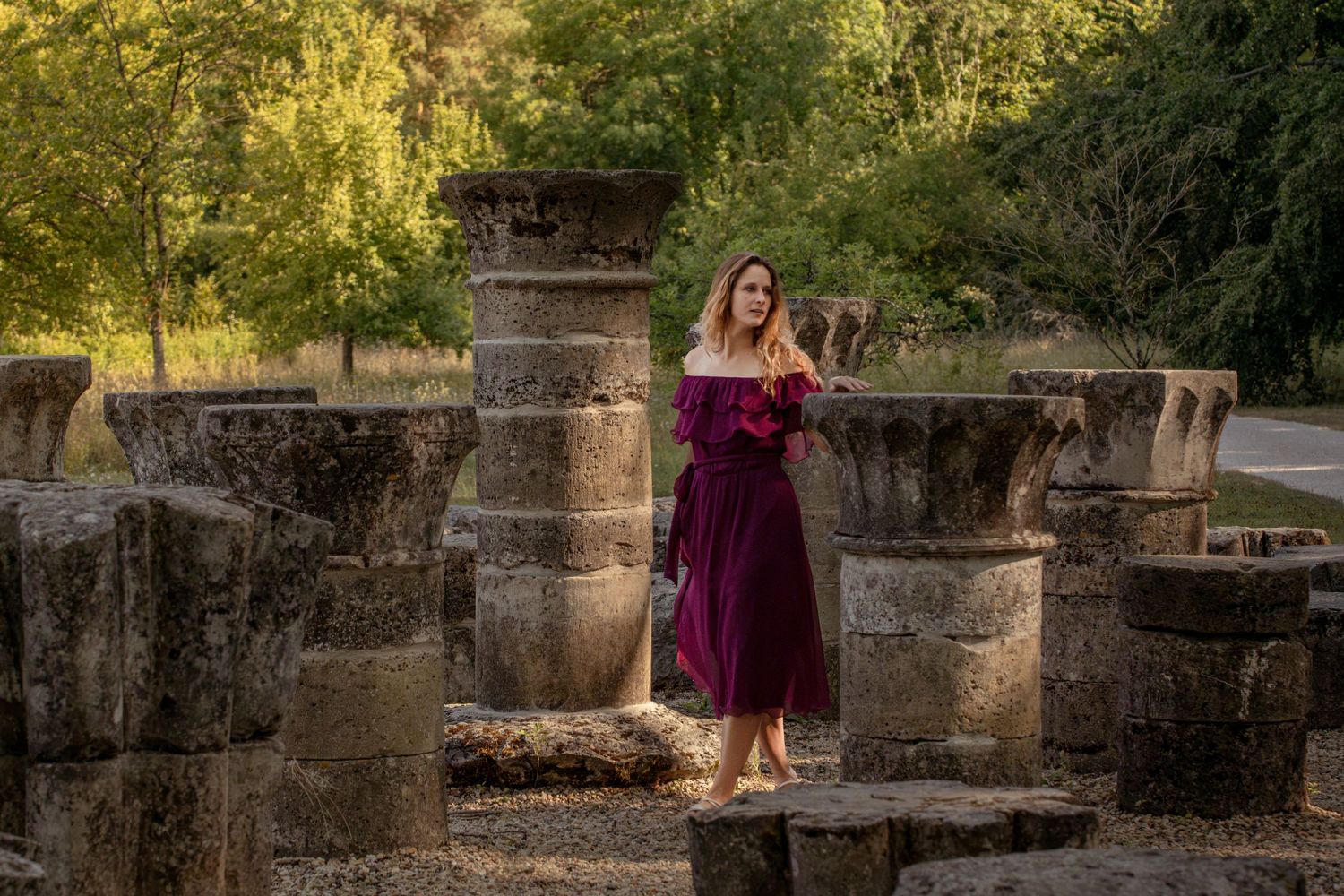Woman in burgundy off-shoulder dress poses among ancient stone ruins surrounded by lush green trees.