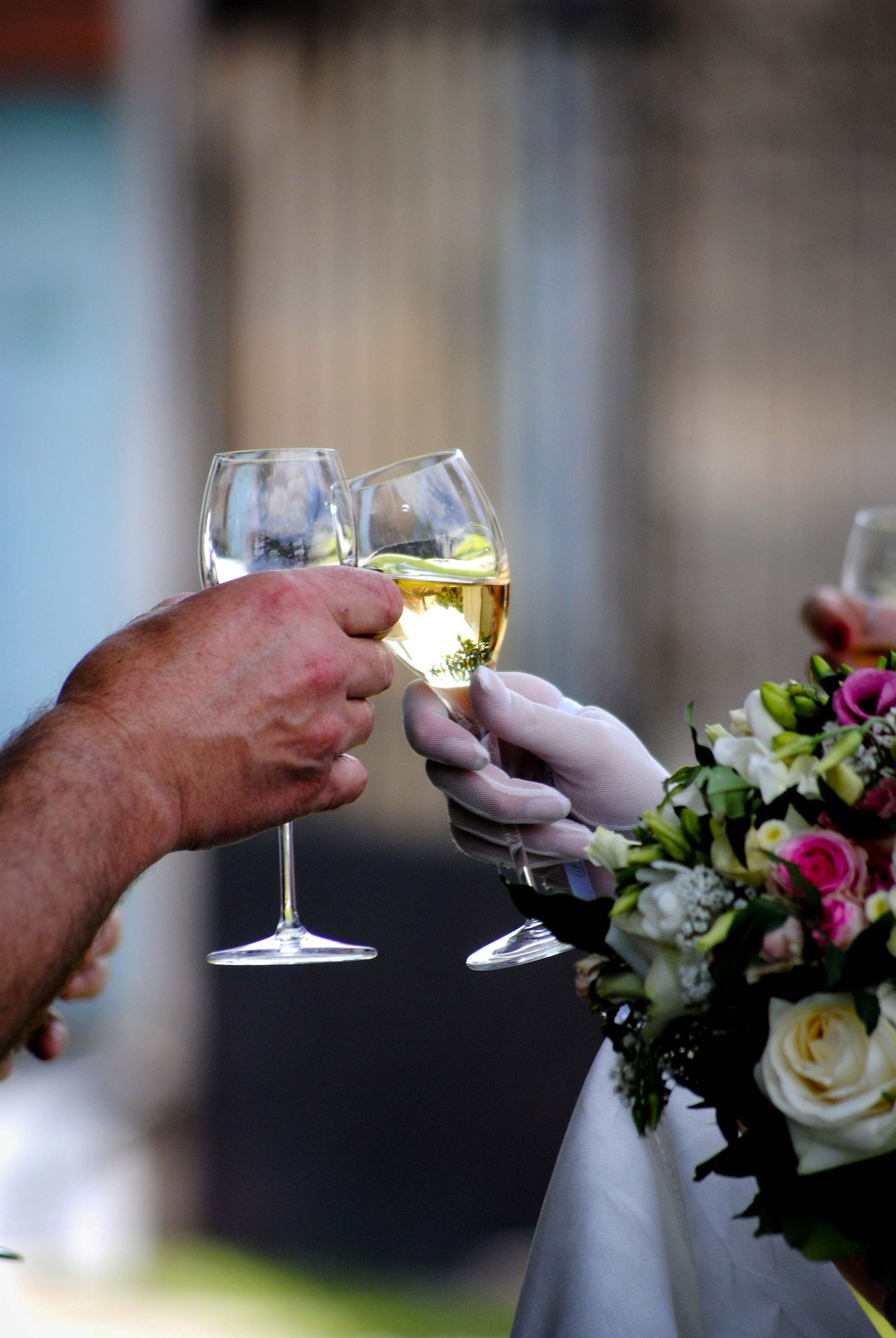 Two hands clinking white wine glasses in a wedding toast, with a bridal bouquet of pink and white flowers nearby.