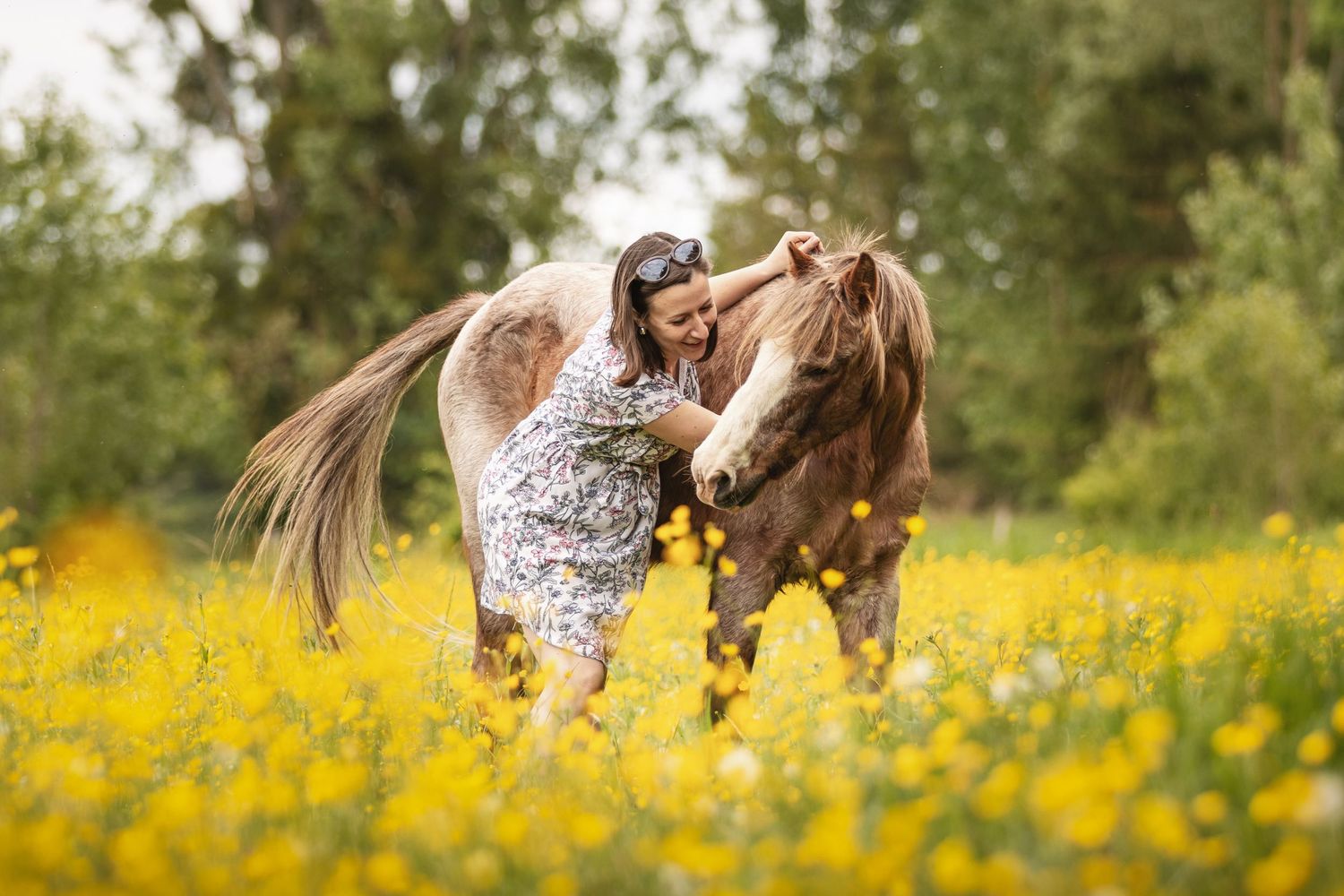 Woman in floral dress affectionately interacting with a brown horse in a vibrant yellow wildflower meadow.