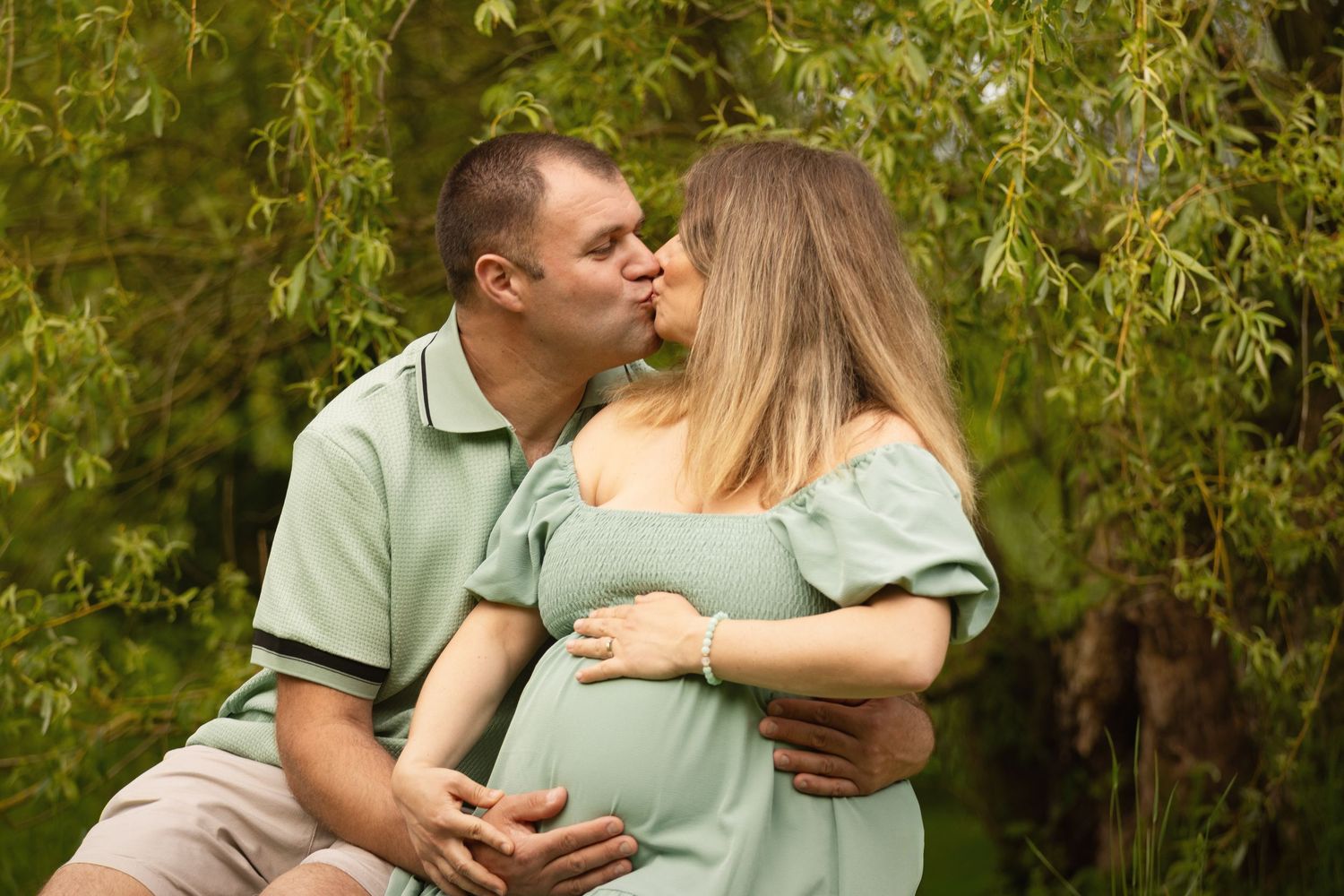 A couple in matching mint green outfits shares a kiss outdoors, with the woman showing a pregnant belly.