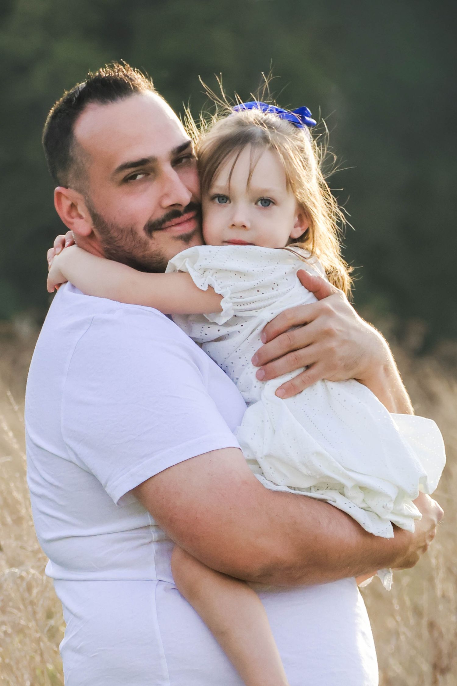 Séance famille lifestyle — papa barbu en chemise blanche serrant tendrement sa fille dans un champ ensoleillé, photographe