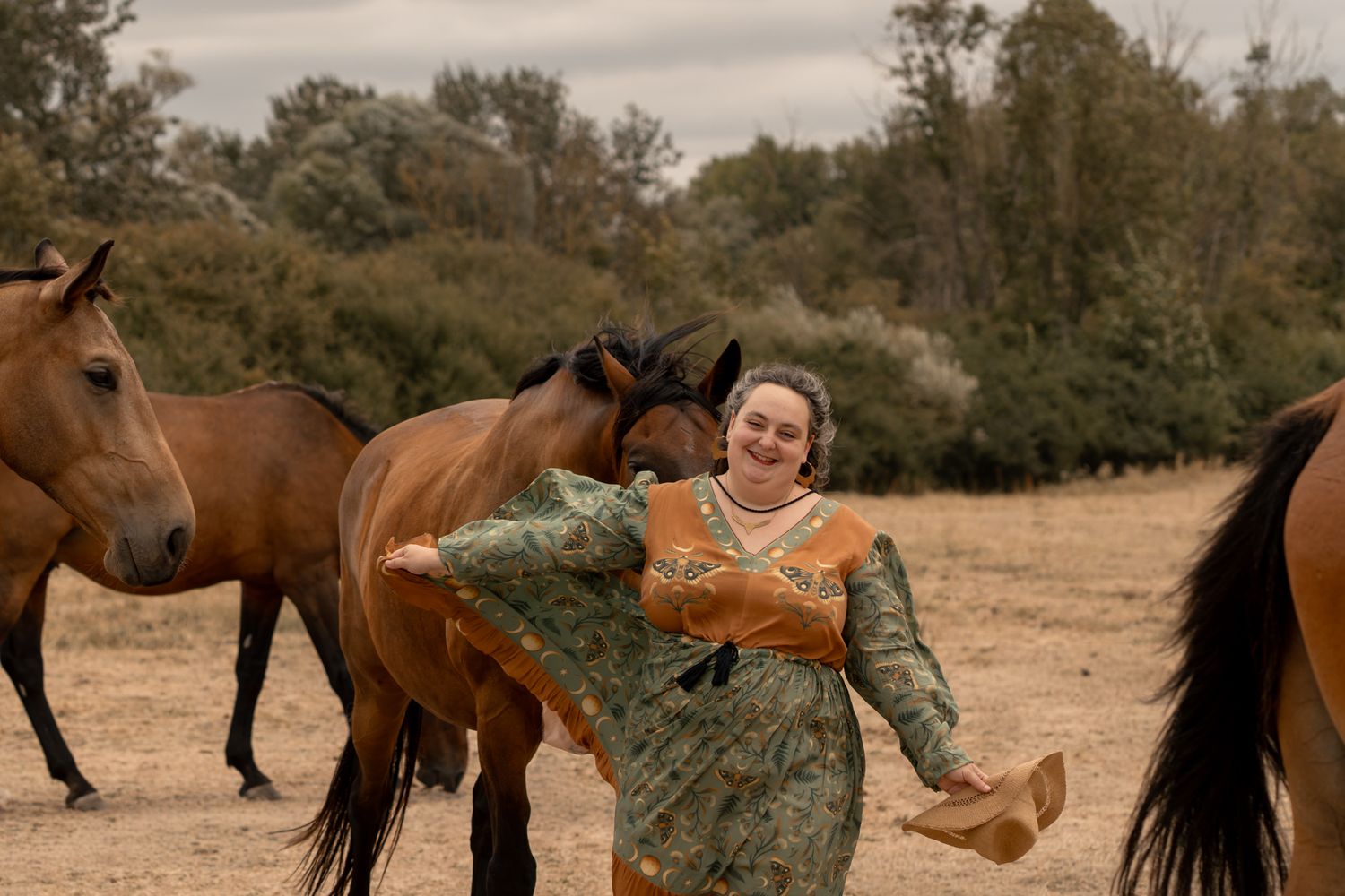 Woman in green floral dress smiling among brown horses on a dry field with trees in the background.
