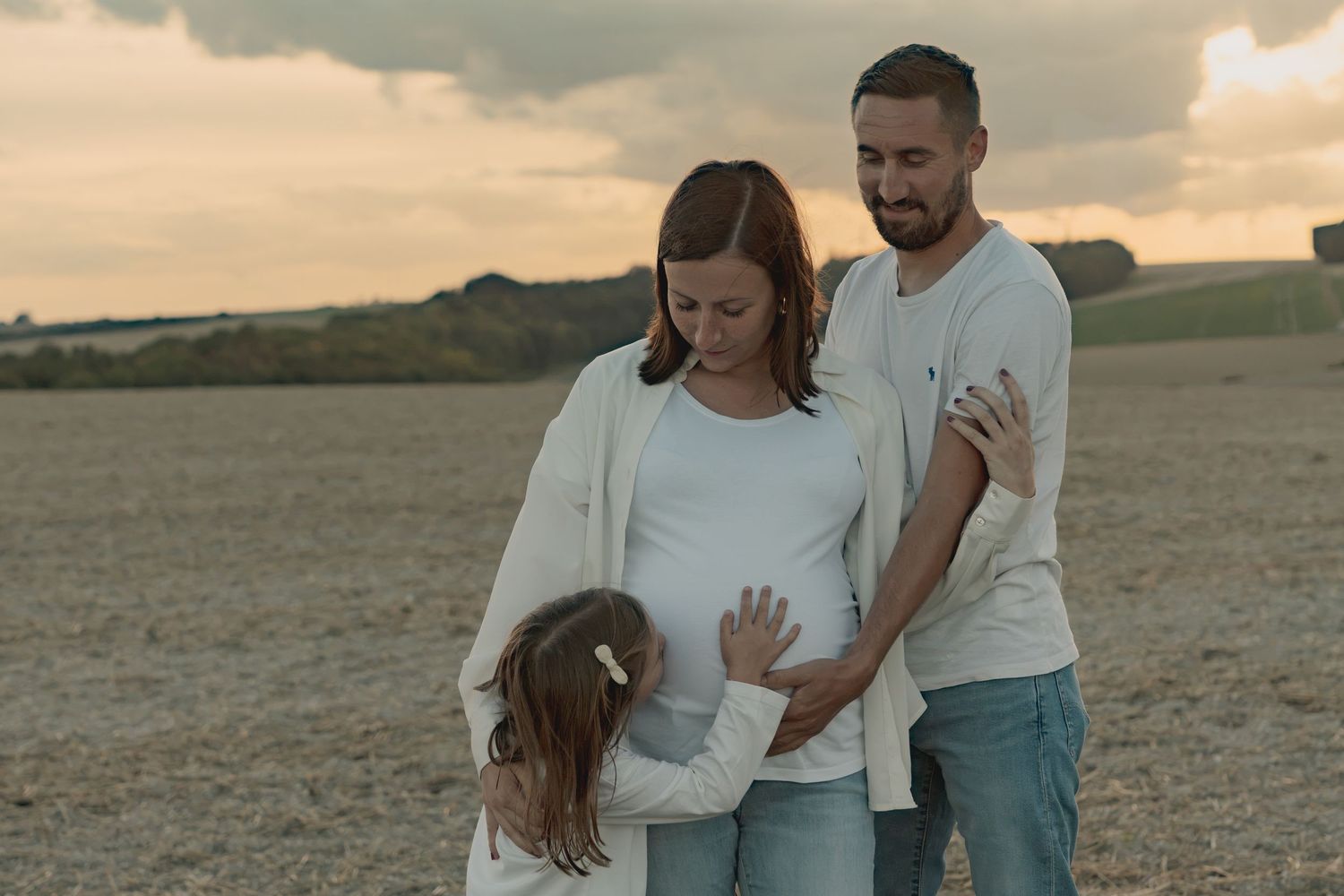 Pregnant woman with husband and daughter touching baby bump in a golden field at sunset.
