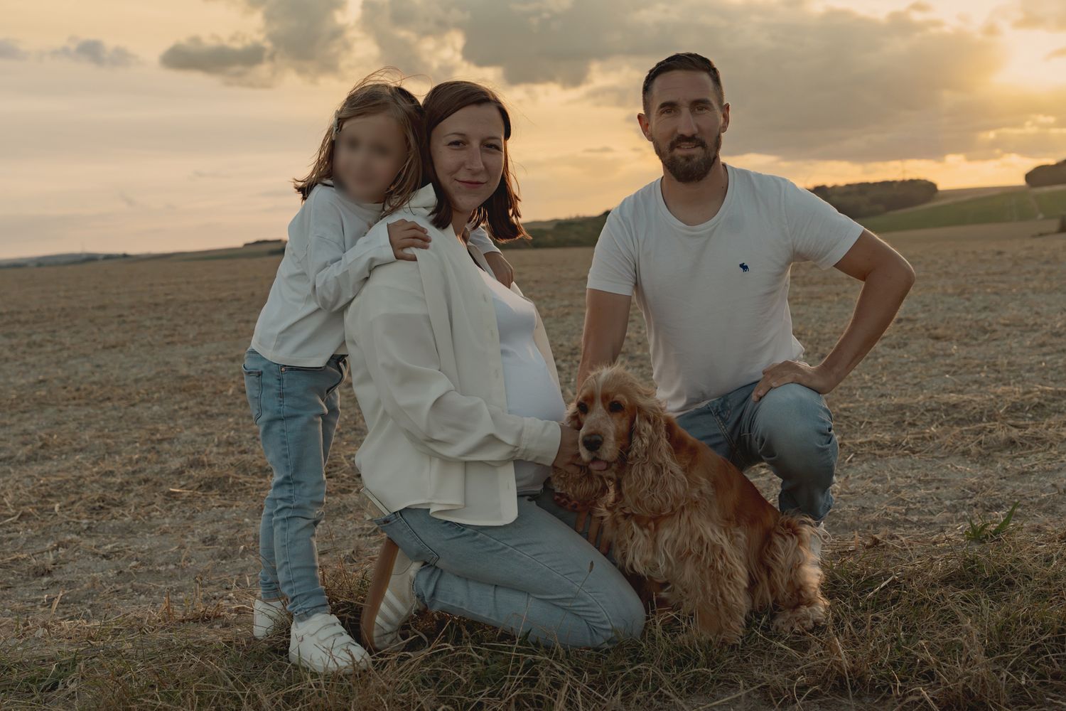 Family photo at sunset in a field with a golden cocker spaniel dog, pregnant woman, man, and young girl.