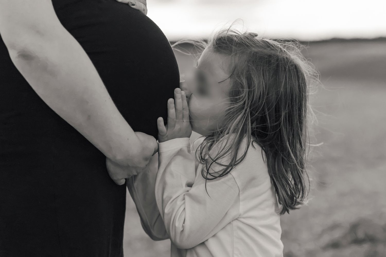 Young girl kissing pregnant mother's belly in tender black and white outdoor maternity photo.