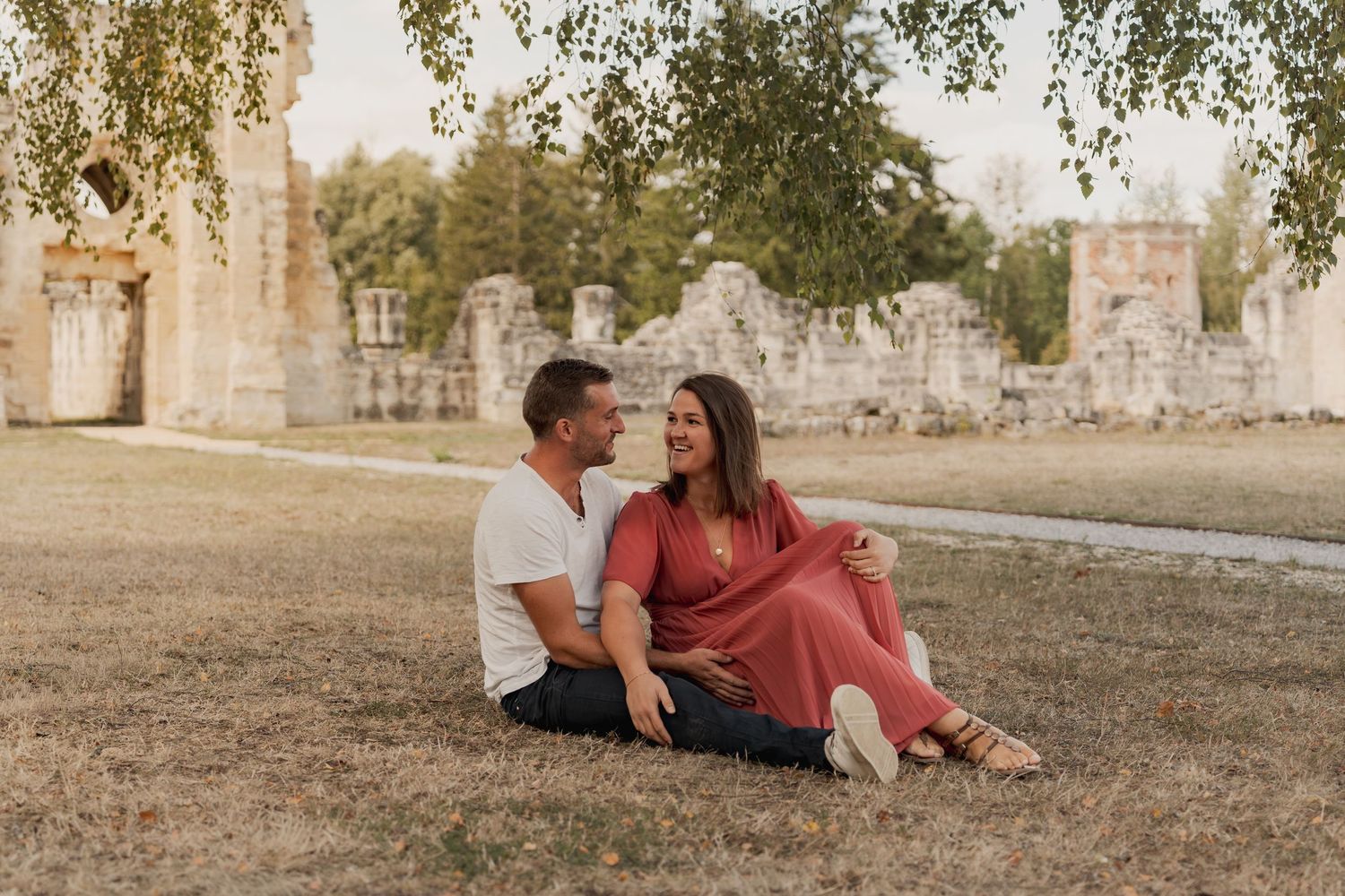 Couple sitting on grass smiling at each other with historic stone ruins and trees in the background.