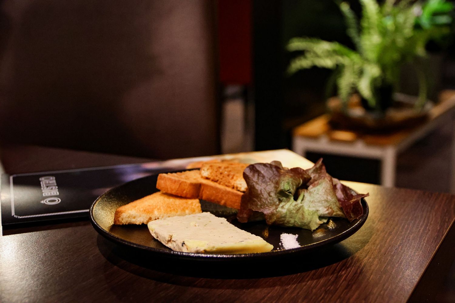 A dark plate with toasted bread slices and cheese served on a wooden table in a dimly lit restaurant.