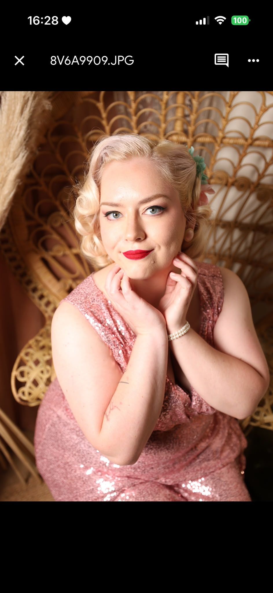 Woman in pink sequin dress with vintage hairstyle and red lips poses in wicker peacock chair for retro glamour portrait.