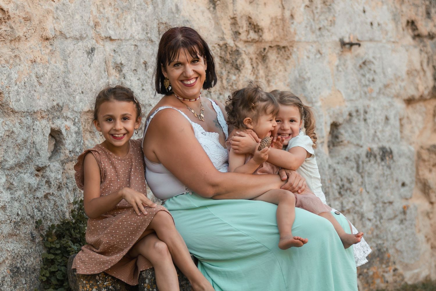 A woman in a mint dress sits with three young girls against a stone wall outdoors.