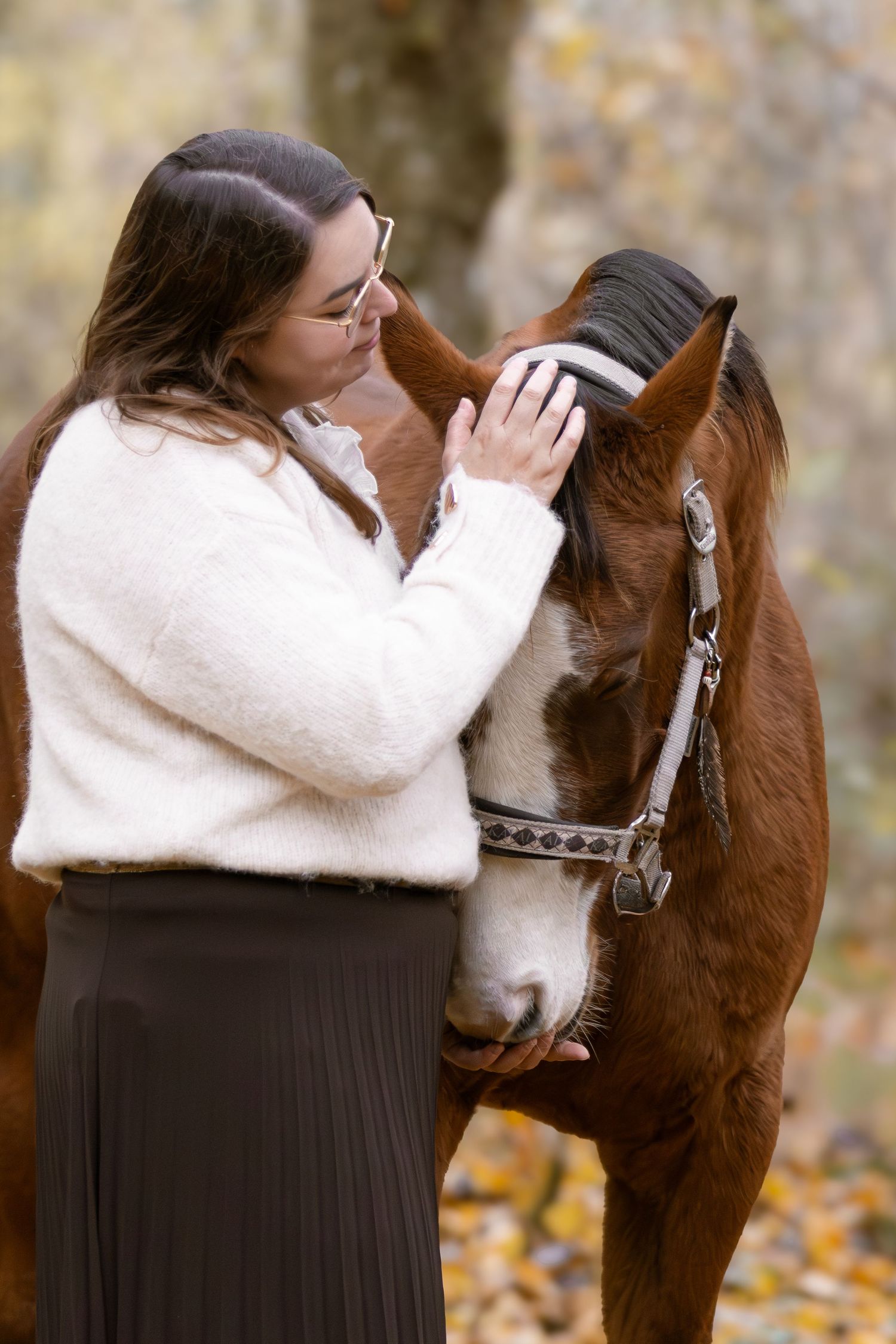 Woman with glasses gently stroking a chestnut horse's forehead in autumn woodland.