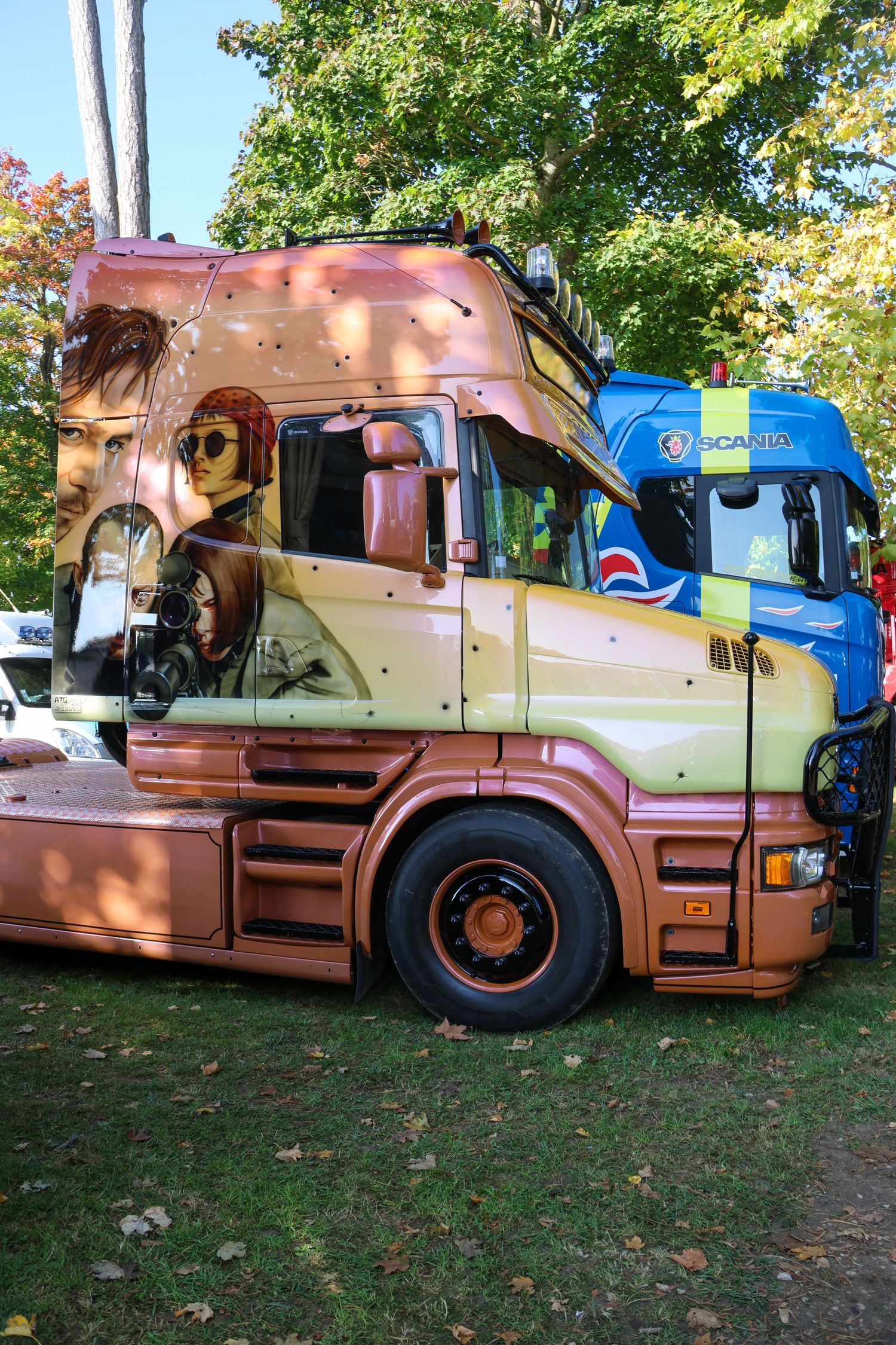 Custom painted Scania truck with bronze finish featuring portrait artwork parked outdoors under trees.