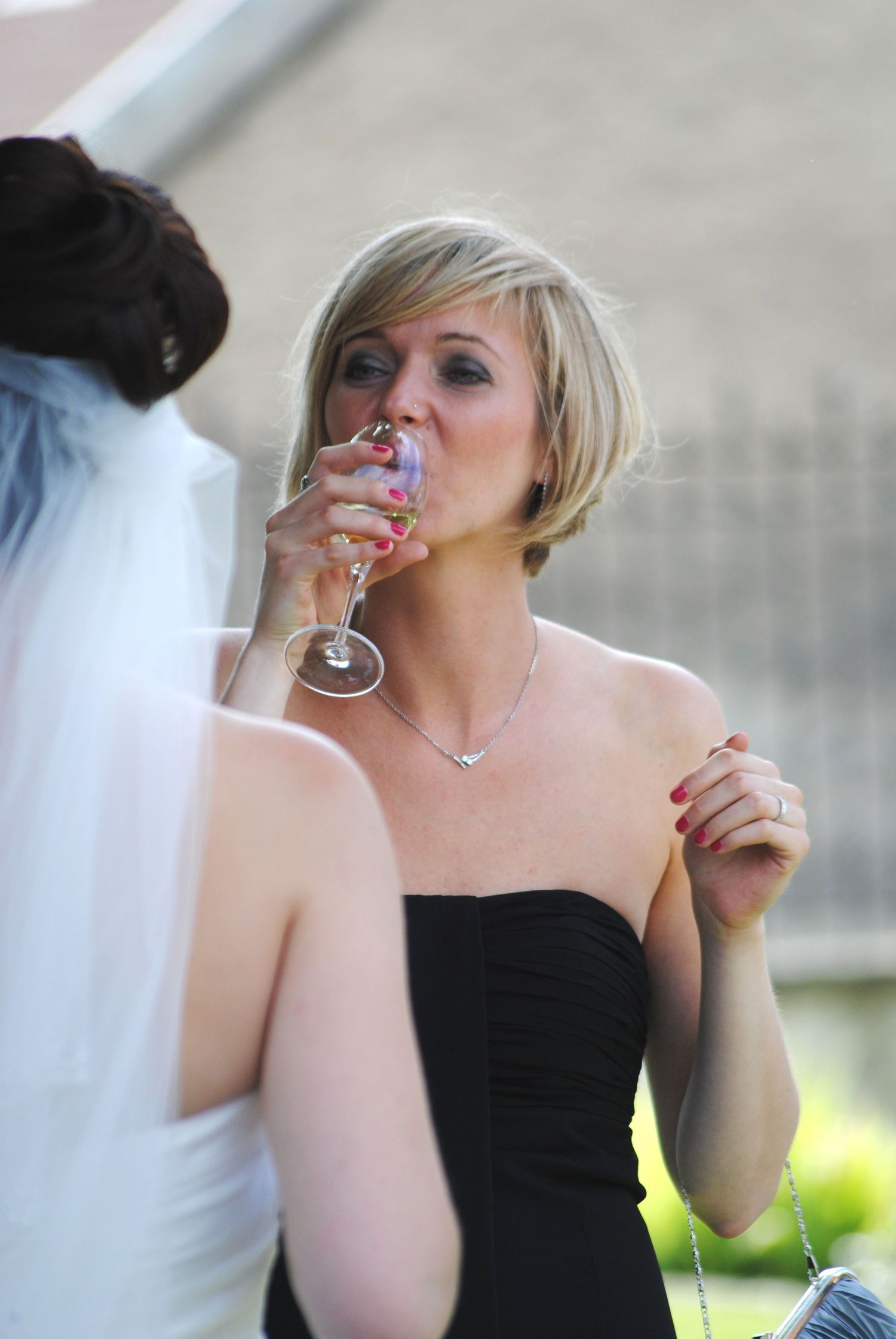 Blonde woman in black strapless dress sipping champagne at a wedding celebration outdoors.