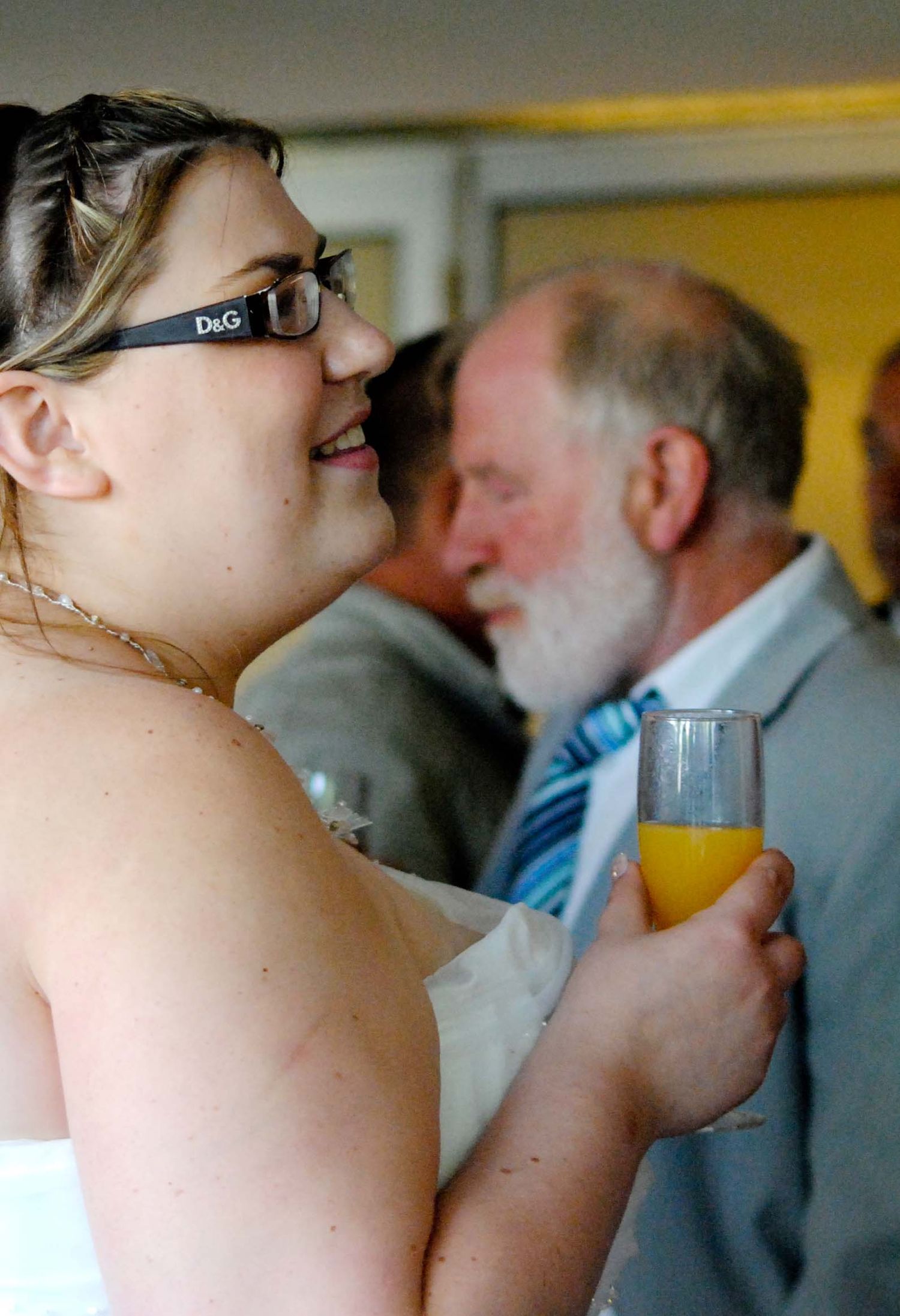 Woman in D&G glasses holding orange juice at a wedding reception with elderly guests in background.