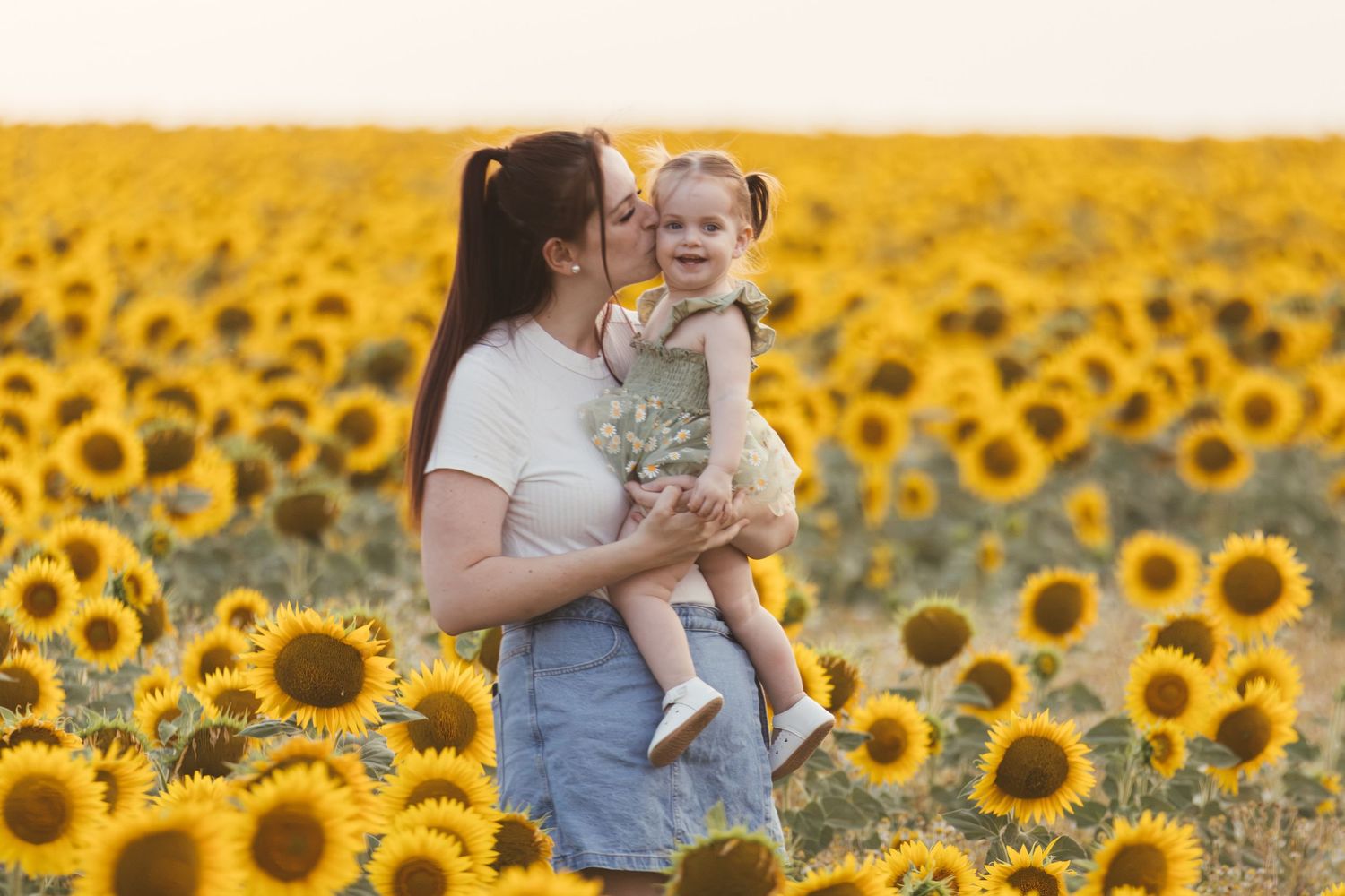 Mother holding smiling toddler daughter in a vibrant sunflower field during golden hour.