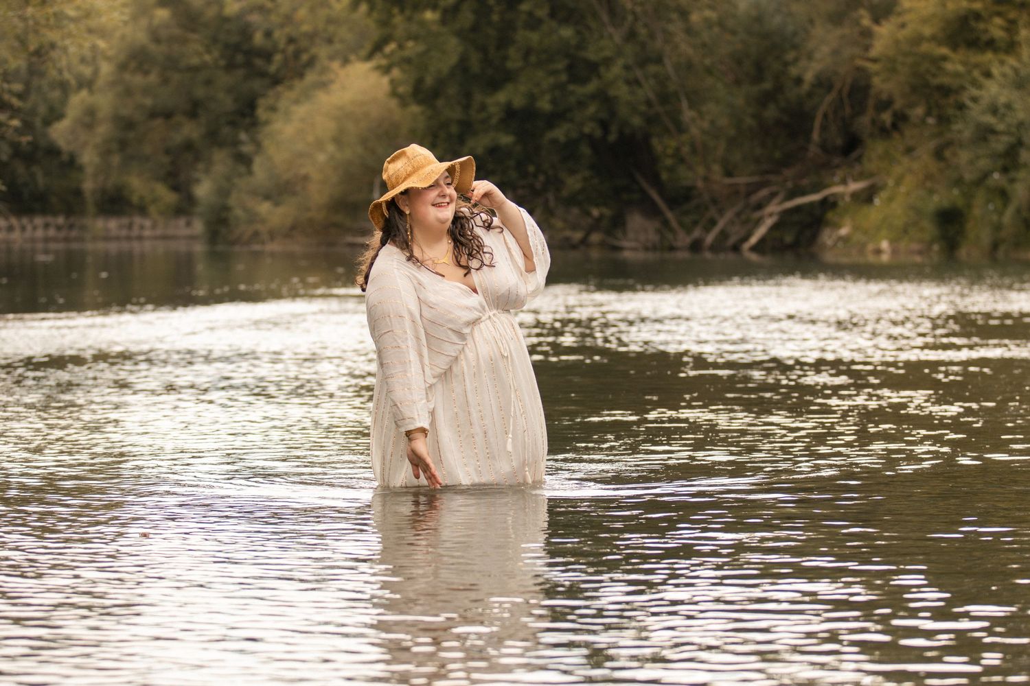 Smiling woman in white dress and straw hat standing in a river surrounded by lush green trees.