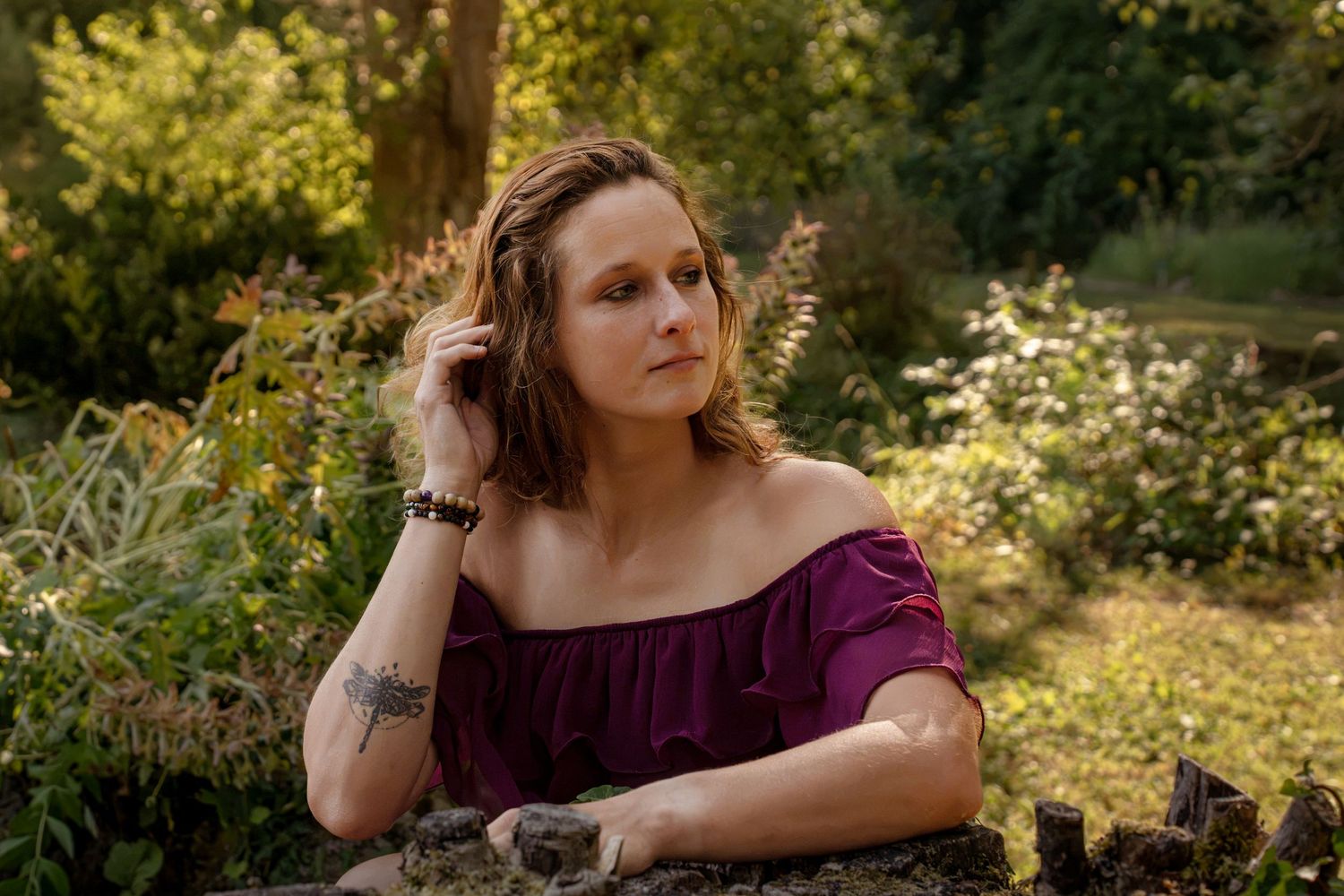 Woman in purple off-shoulder top poses thoughtfully in lush green garden setting with tattoos visible.