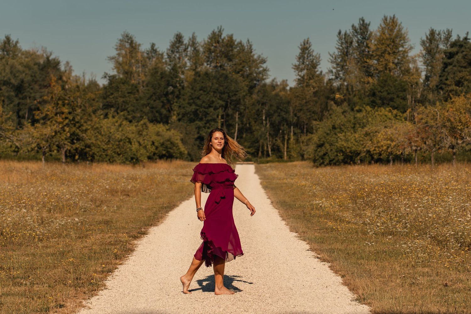 Woman in burgundy off-shoulder dress walking on gravel path surrounded by green trees on a sunny day.