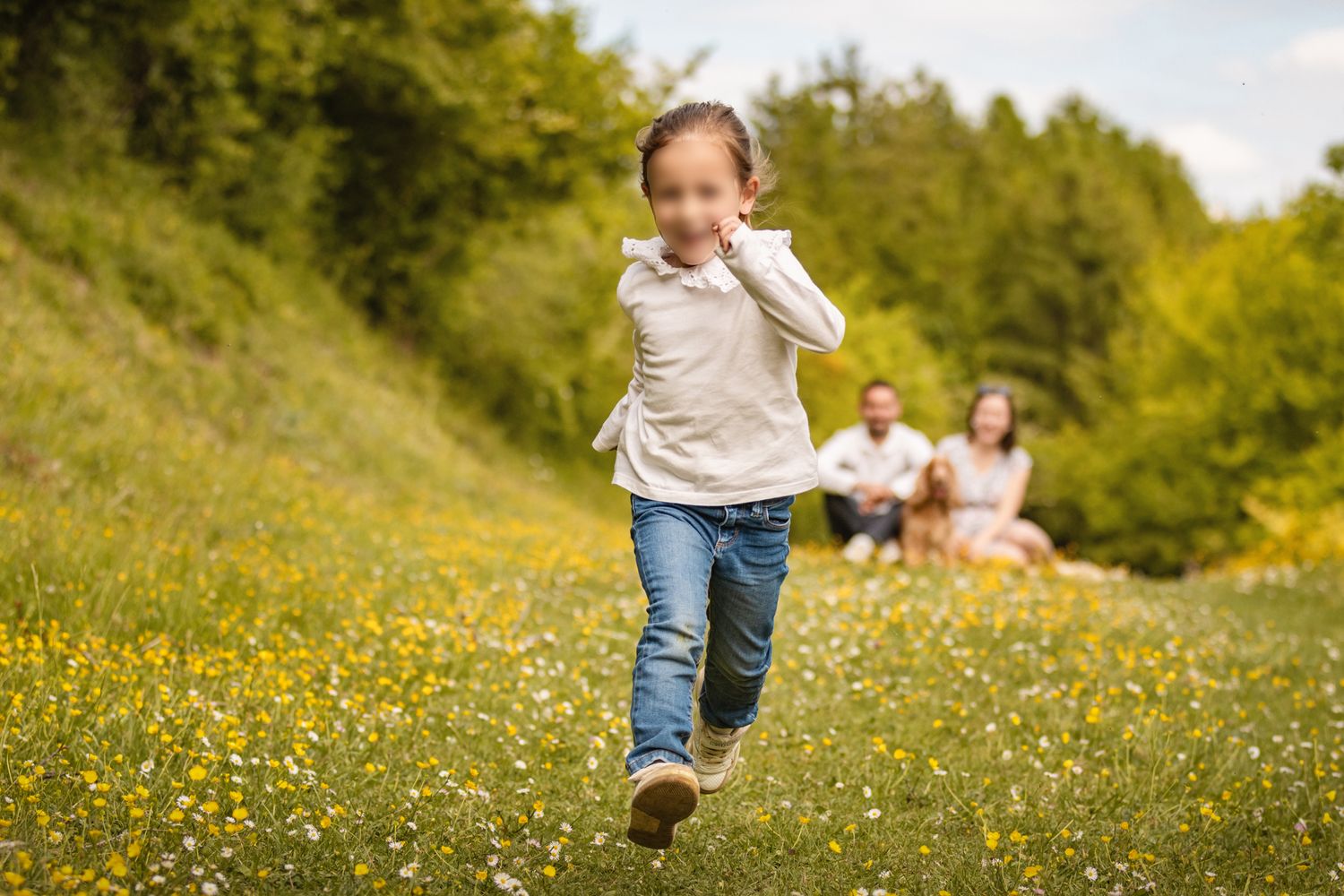 Young girl running through sunny wildflower meadow with parents relaxing in background on green hillside.