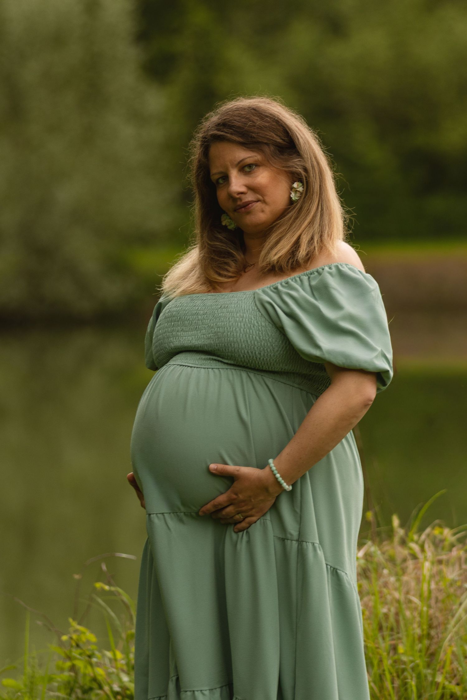 Pregnant woman in sage green off-shoulder dress cradles baby bump outdoors near a serene lake.