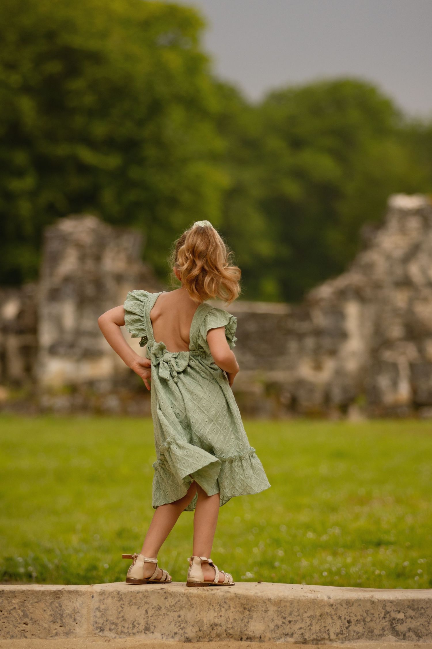 Young girl in green backless dress with ruffles stands on stone ledge facing ancient ruins in lush garden.