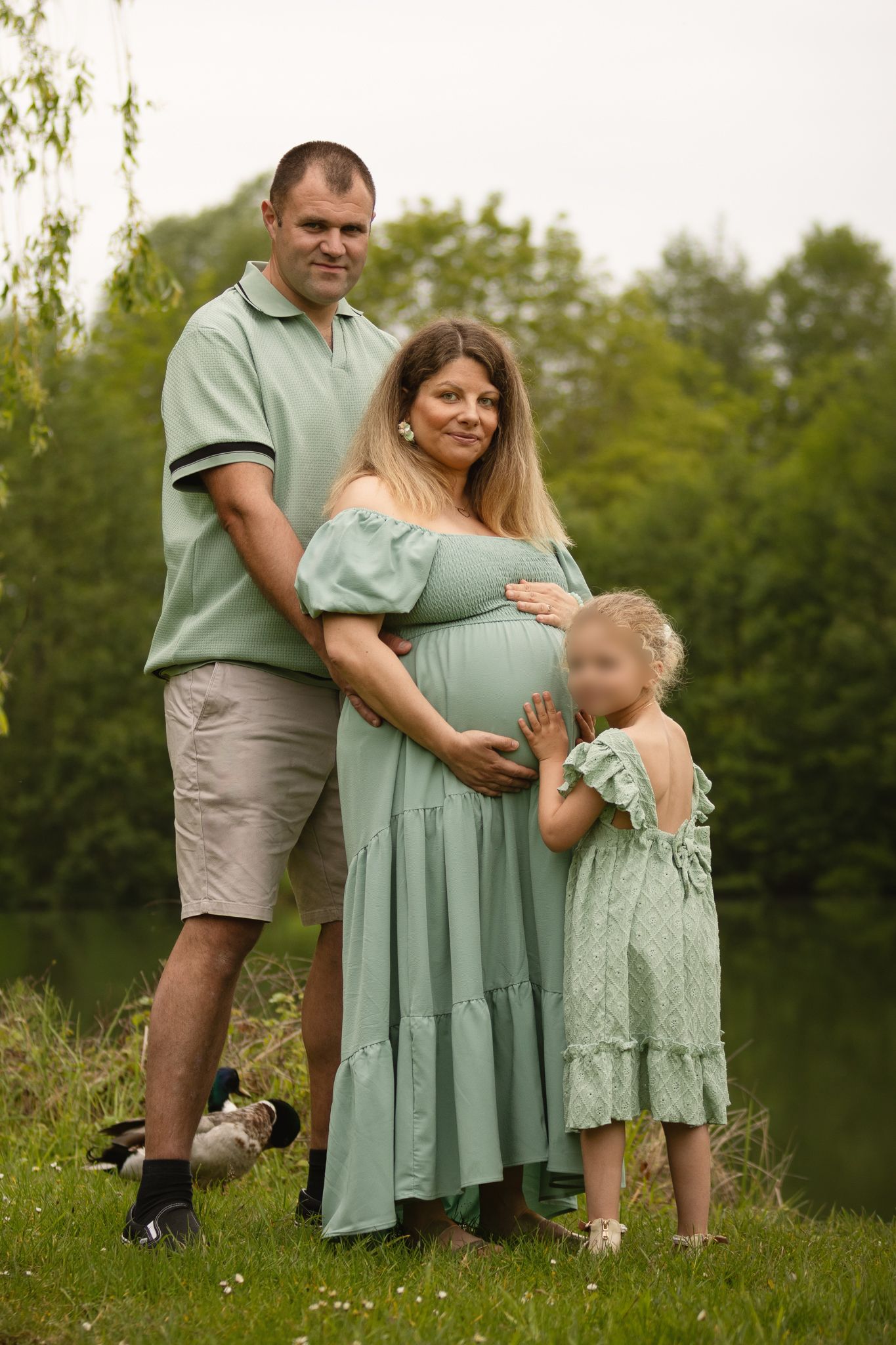 Pregnant woman in green dress poses with husband and young daughter outdoors near a pond surrounded by trees.