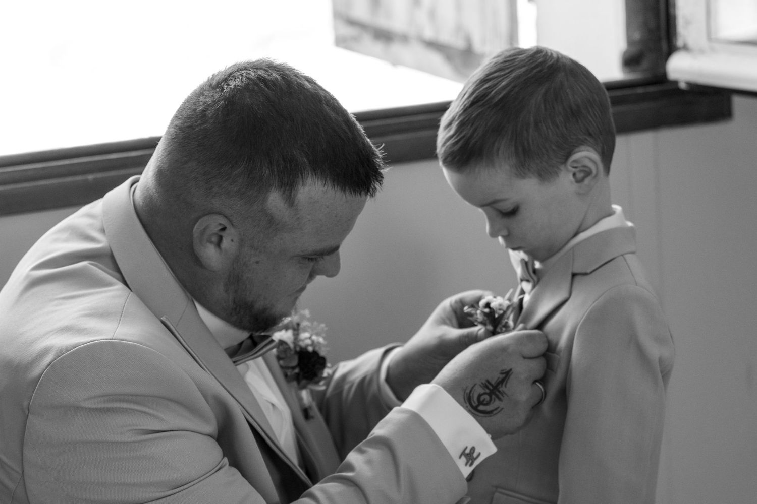Black and white photo of a man in a suit pinning a boutonniere on a young boy in a suit at a wedding.