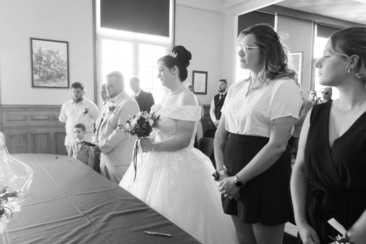 Black and white wedding photo of bride in white gown holding bouquet standing with guests in elegant venue.