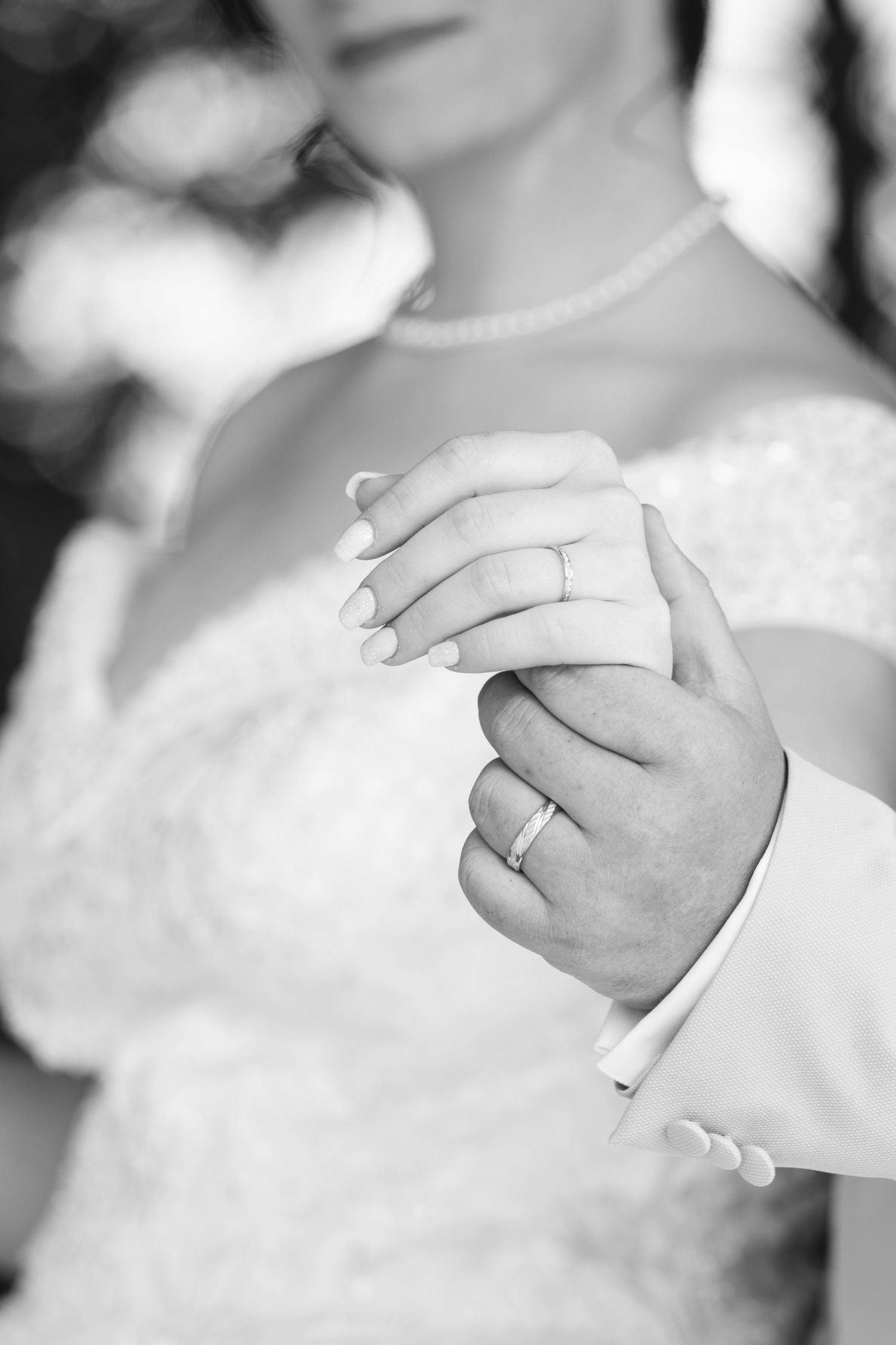 Black and white close-up of newlyweds holding hands showing wedding rings, bride in lace dress.