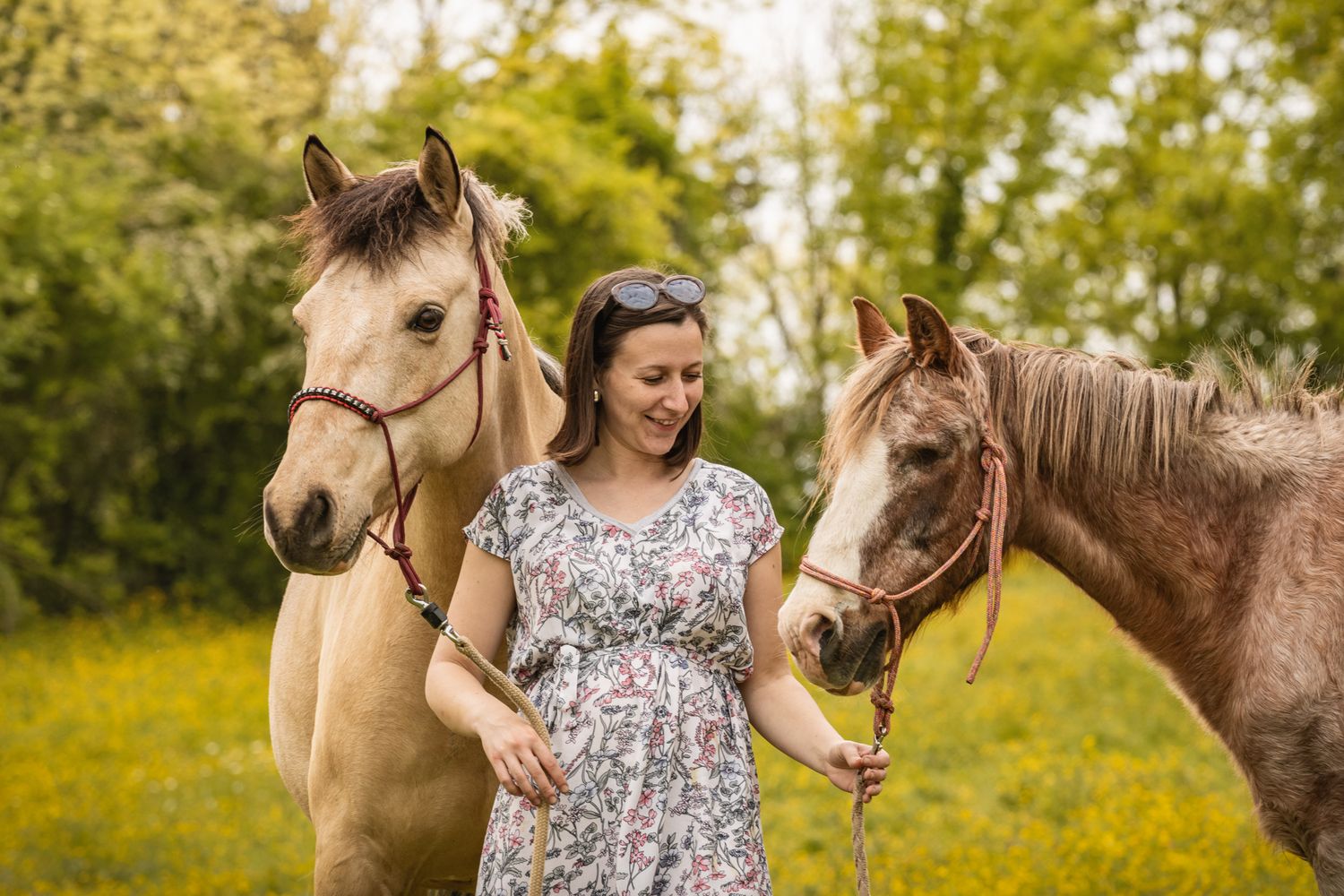 Pregnant woman in floral dress smiling while standing between two brown horses in a green meadow.