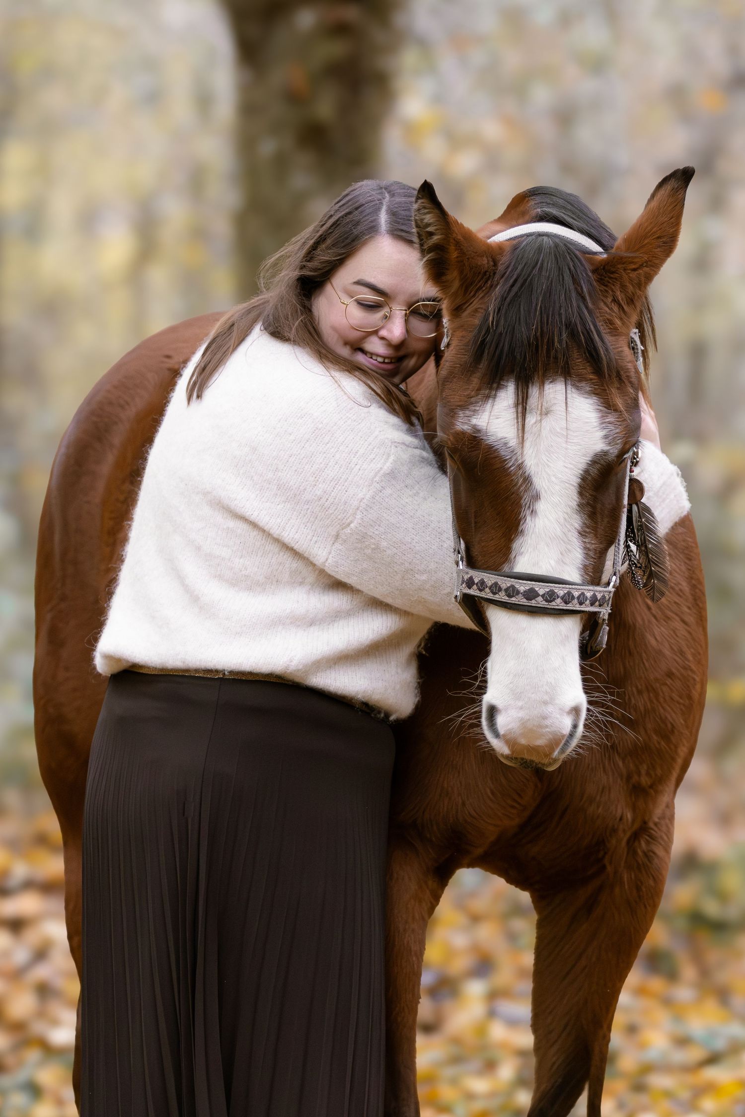 Woman in white sweater embracing a brown and white horse in an autumn forest setting.