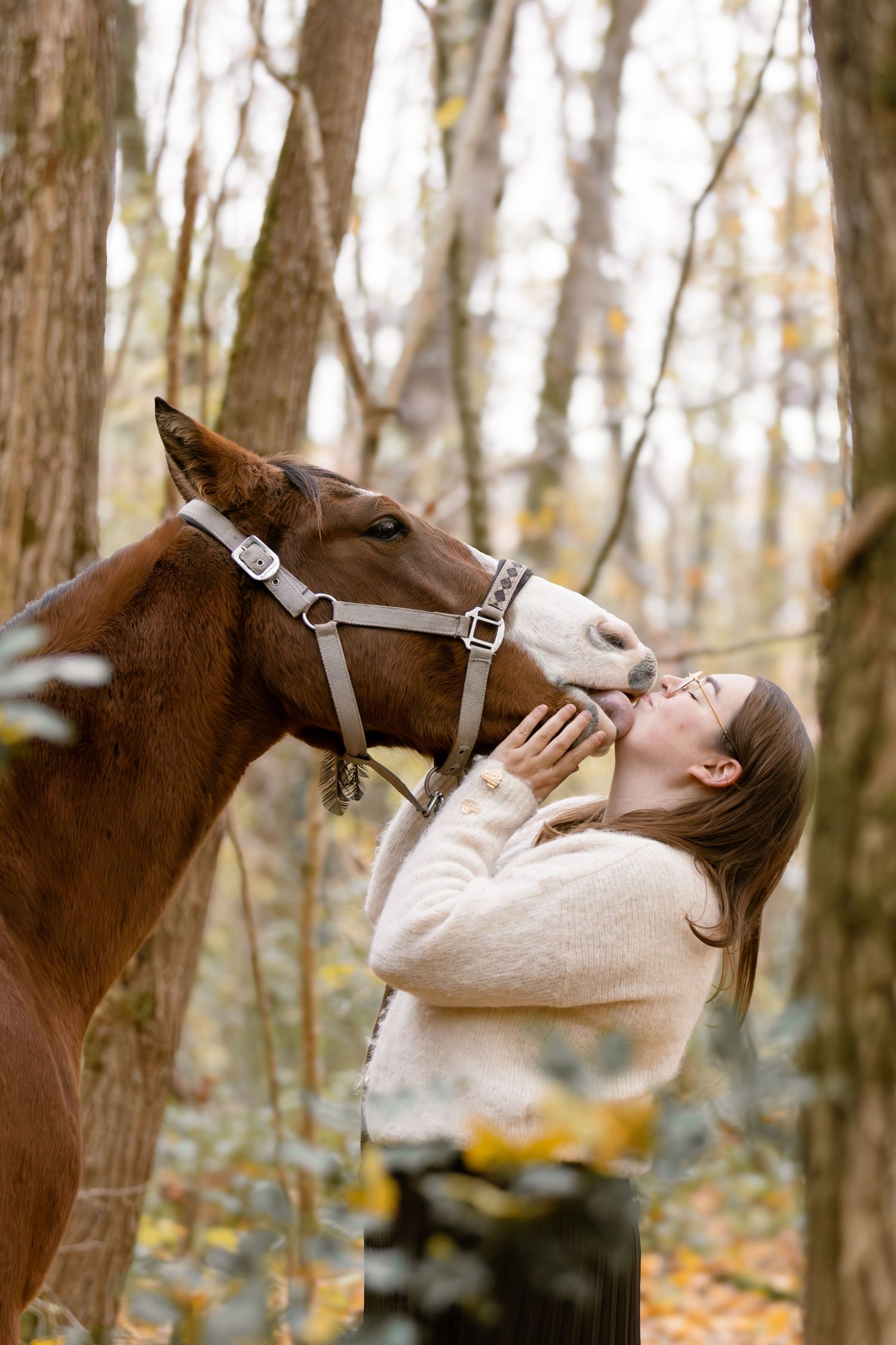 Young woman in cream sweater sharing a tender kiss with a brown horse in autumn woods.