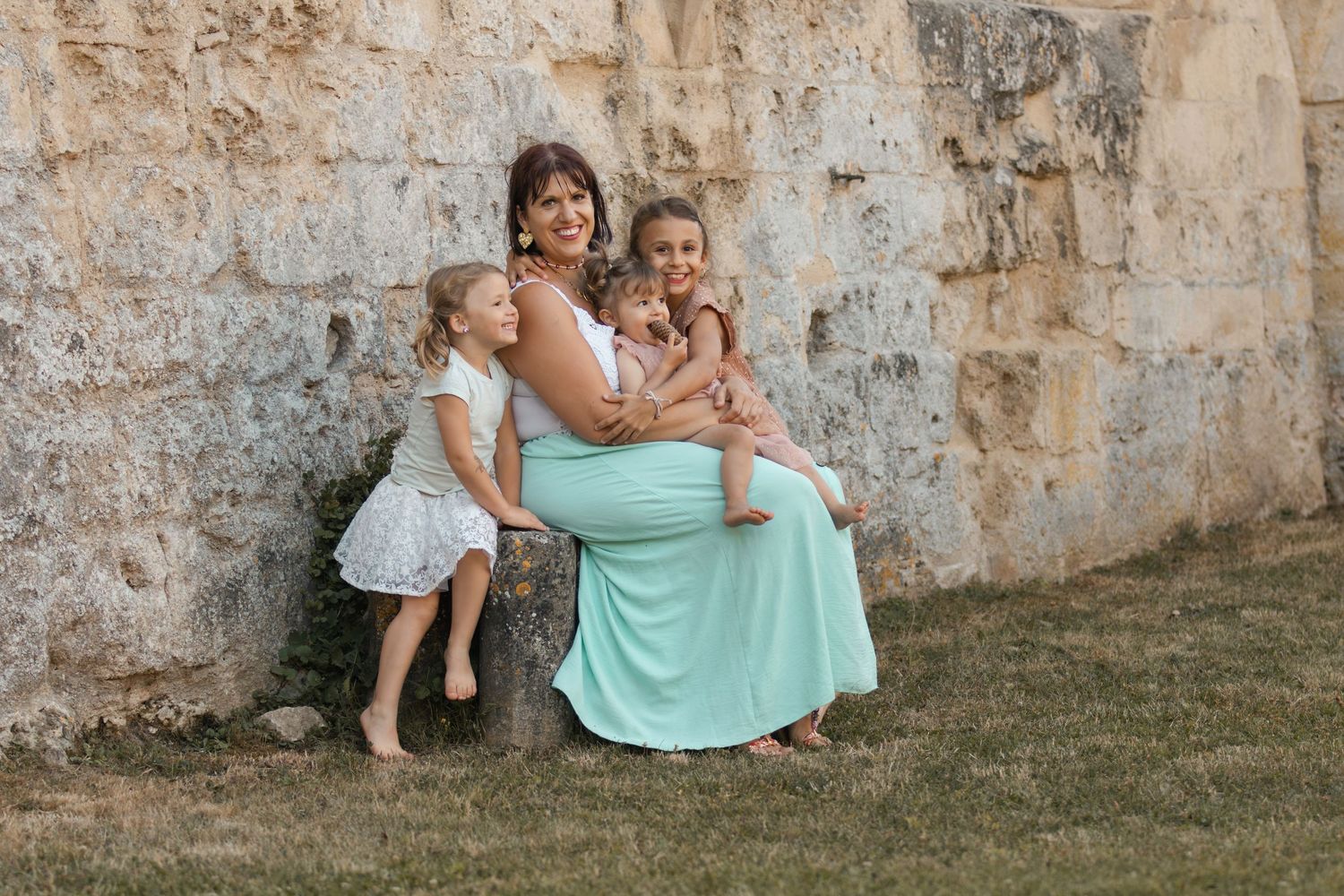 Pregnant woman in mint maxi dress sits with two young daughters against rustic stone wall outdoors.
