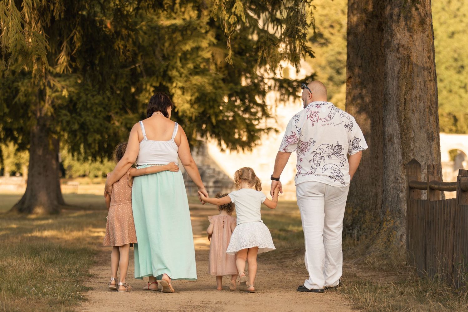 Family of four walking hand-in-hand along a sunny tree-lined path in a park.