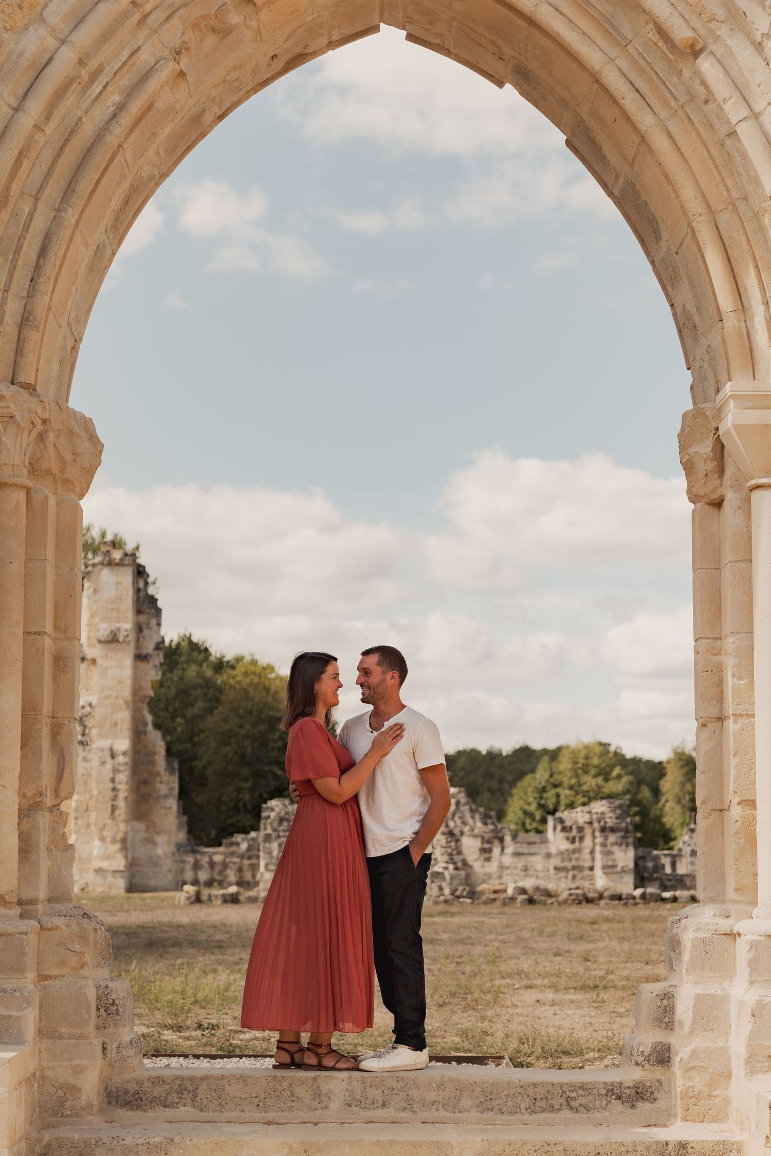 Couple standing under a stone arch at ancient ruins, woman in red dress and man in white shirt gazing at each other.