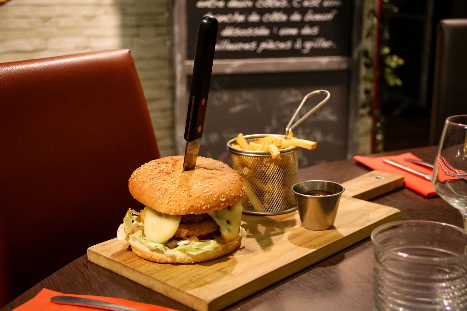 A gourmet burger with a knife stuck in it served on a wooden board with crispy fries in a metal cup at a restaurant.