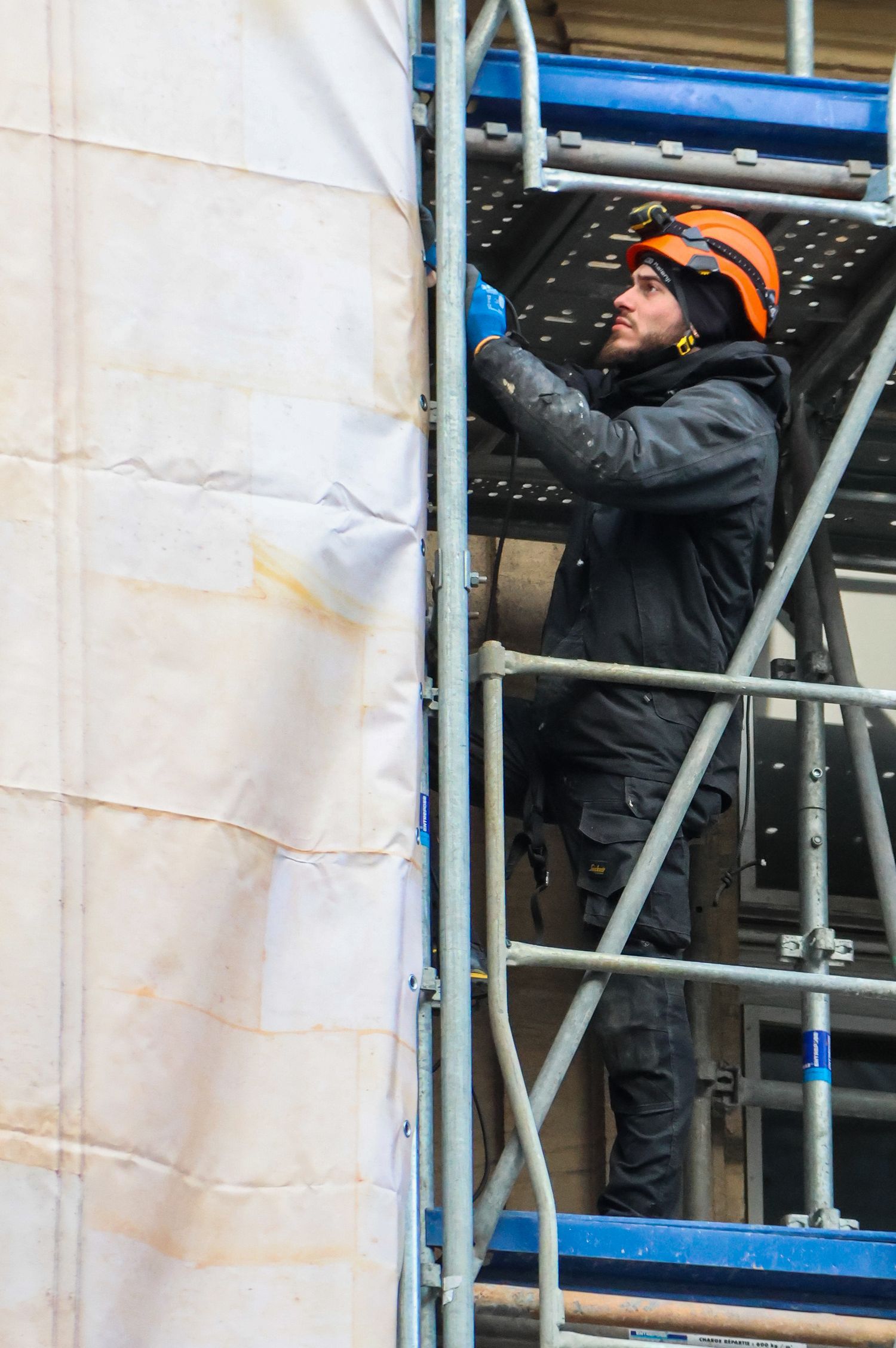 Construction worker in orange helmet on scaffolding inspecting building exterior during renovation work.