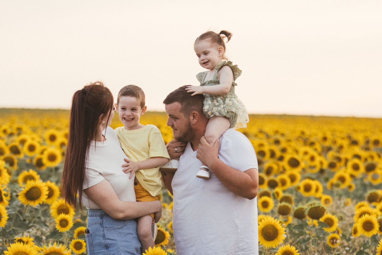 Happy family with two young children posing in a vibrant sunflower field at golden hour sunset.