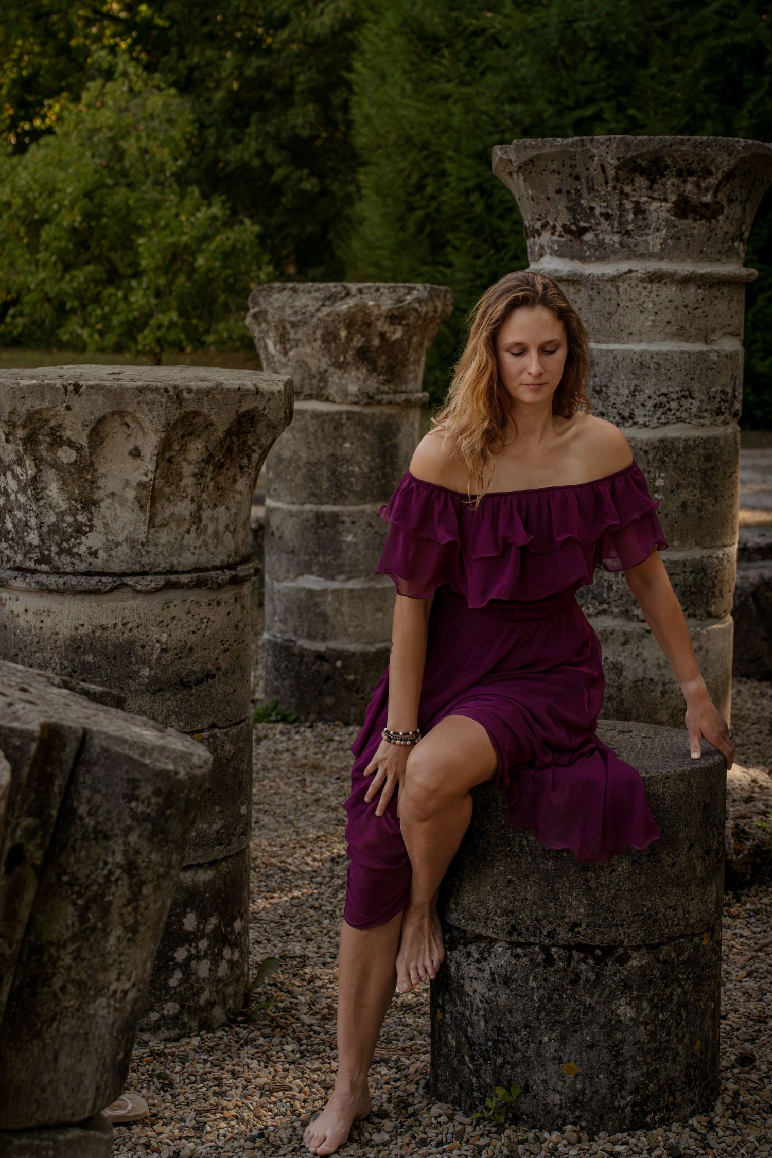 Woman in purple off-shoulder dress sitting on ancient stone columns surrounded by lush greenery.