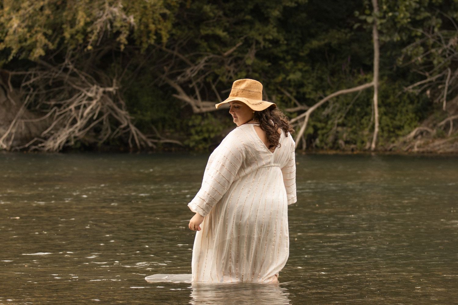 Pregnant woman in white dress and tan hat standing in a serene river with lush green trees in background.