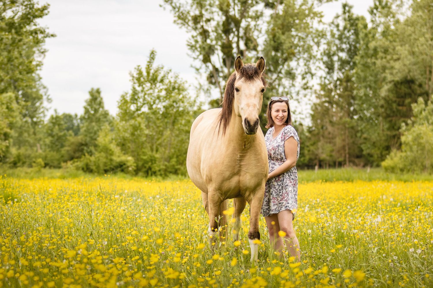 Woman smiling beside a palomino horse in a sunny yellow wildflower meadow with green trees in background.