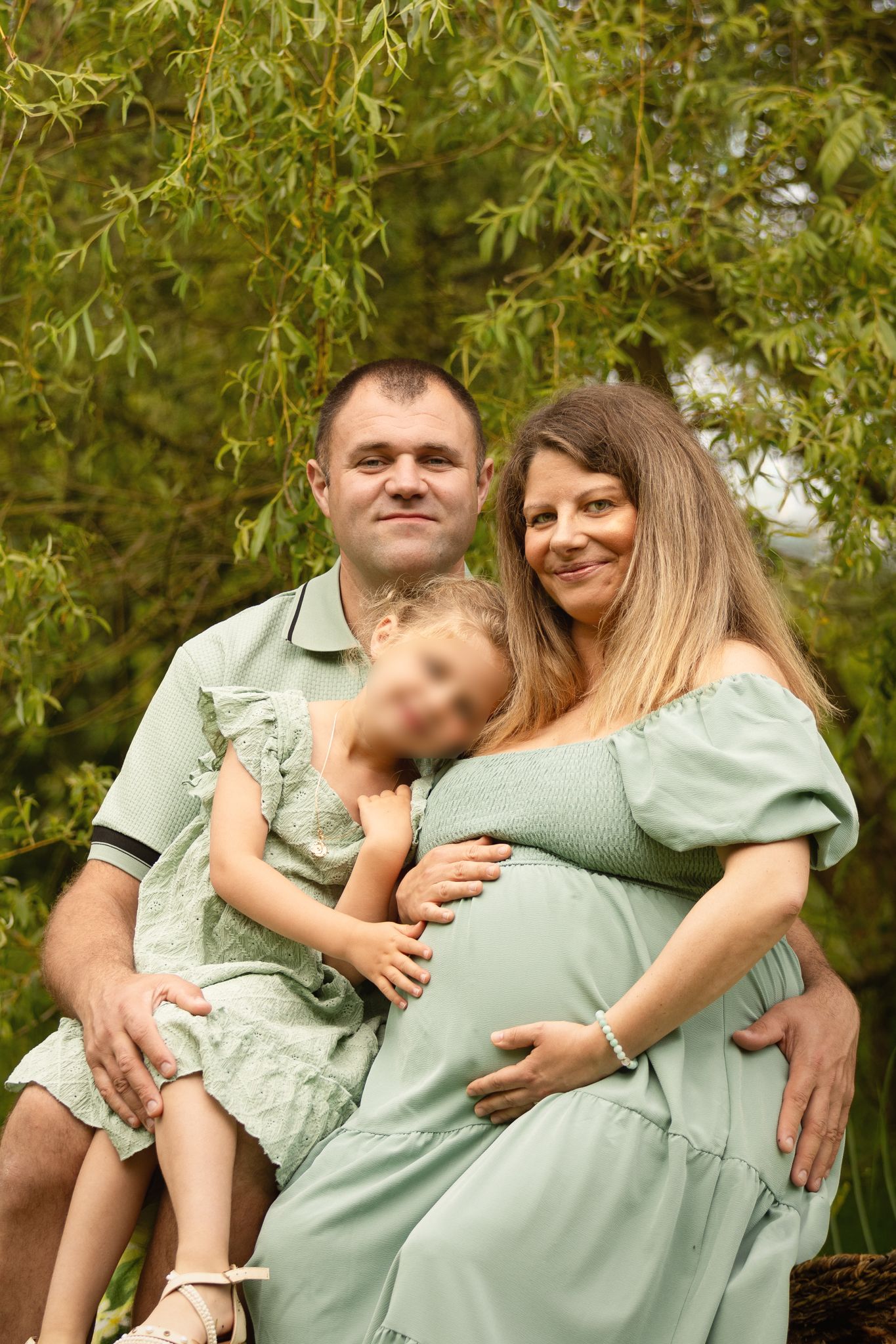 Happy family with pregnant mother, father, and young child posing outdoors among lush green willow trees.