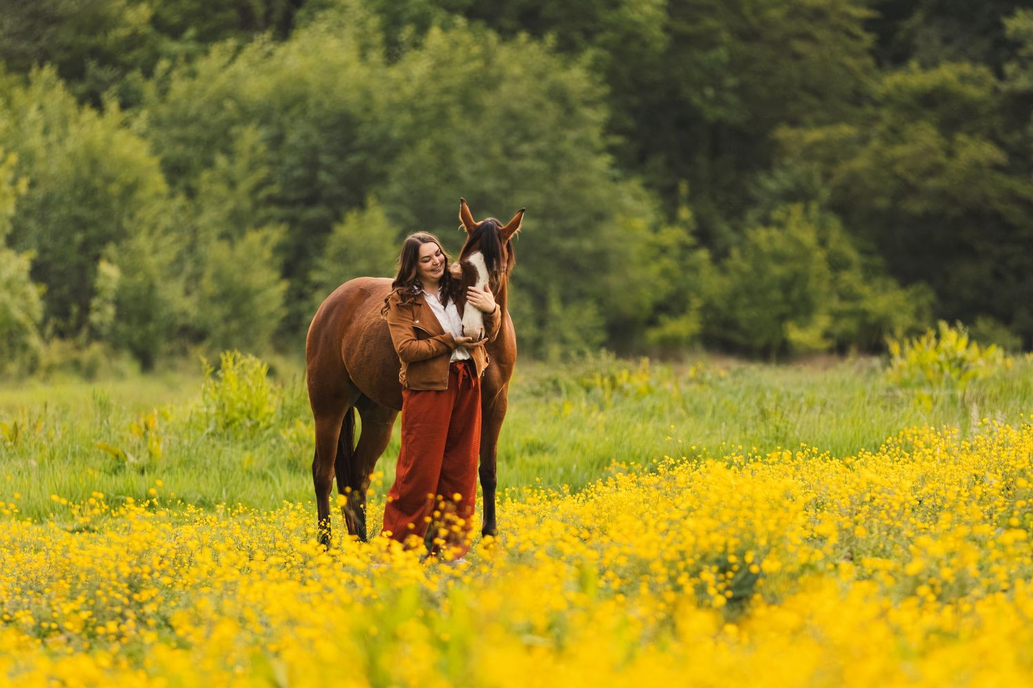 Woman in orange dress standing with brown horse in yellow wildflower meadow with green trees background.