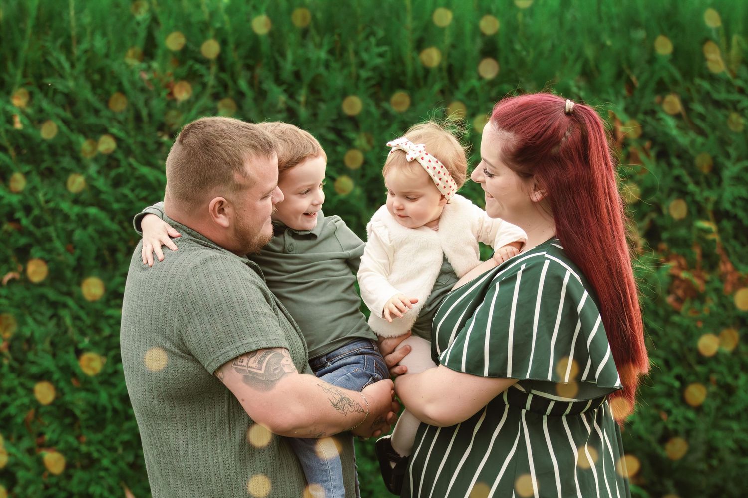 Happy family of four with two young children embracing in front of lush green floral backdrop.