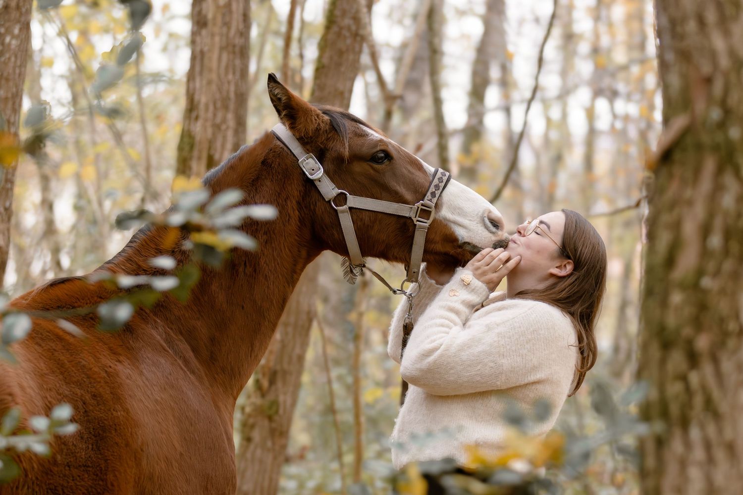A laughing woman in a cream sweater receives a gentle nuzzle from a chestnut horse in the woods.