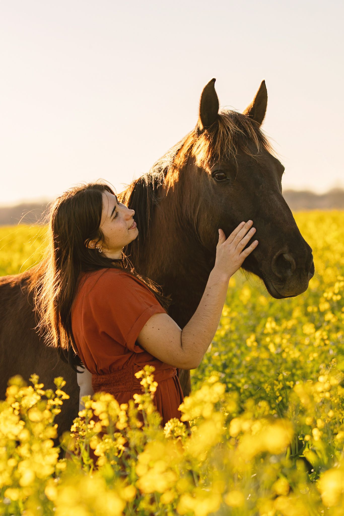 Woman in red dress gently petting a dark horse in a vibrant yellow flower field during golden hour.