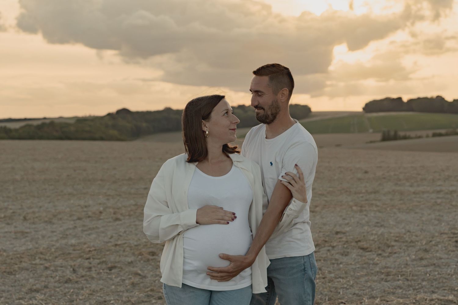 Pregnant couple embracing in a golden field at sunset, sharing a tender moment outdoors.