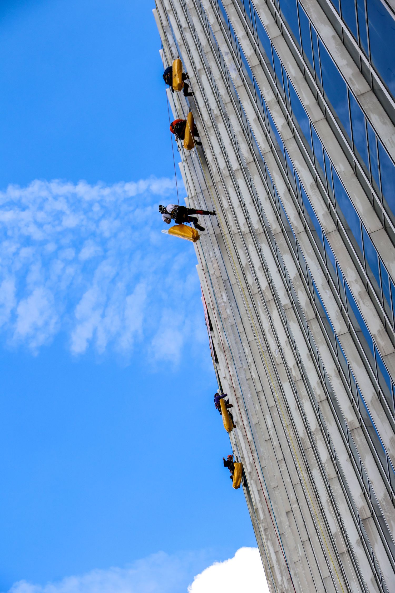 Window washers suspended on yellow scaffolding clean a tall glass skyscraper facade against a bright blue sky.