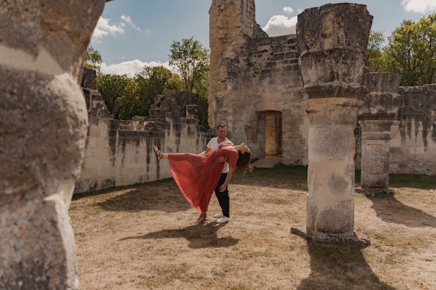 Couple dancing romantically among ancient stone ruins, woman in flowing red dress, surrounded by columns and lush trees.
